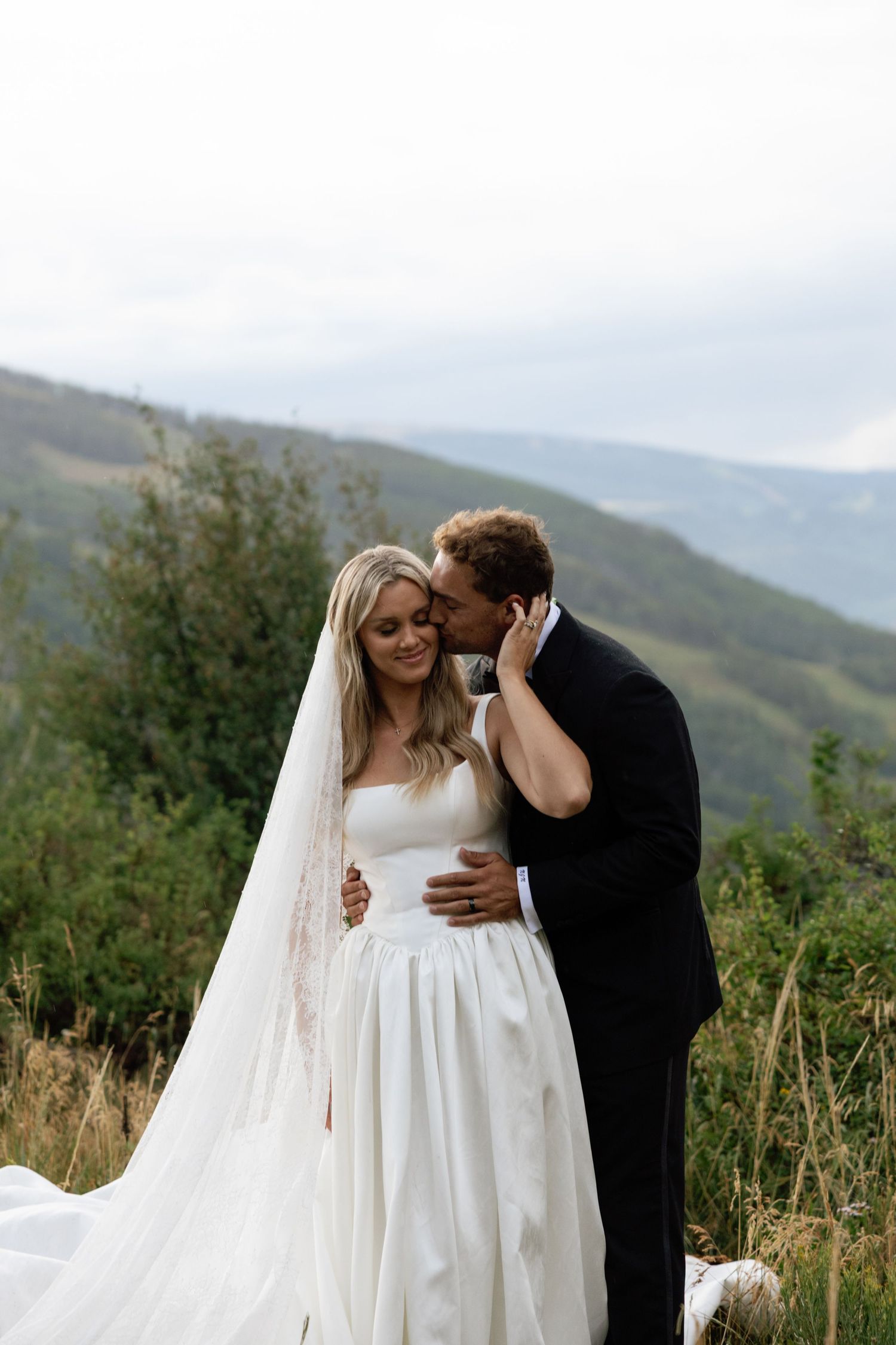 A bride and groom share a romantic moment on a mountain overlook, with the bride in a flowing white gown and veil.