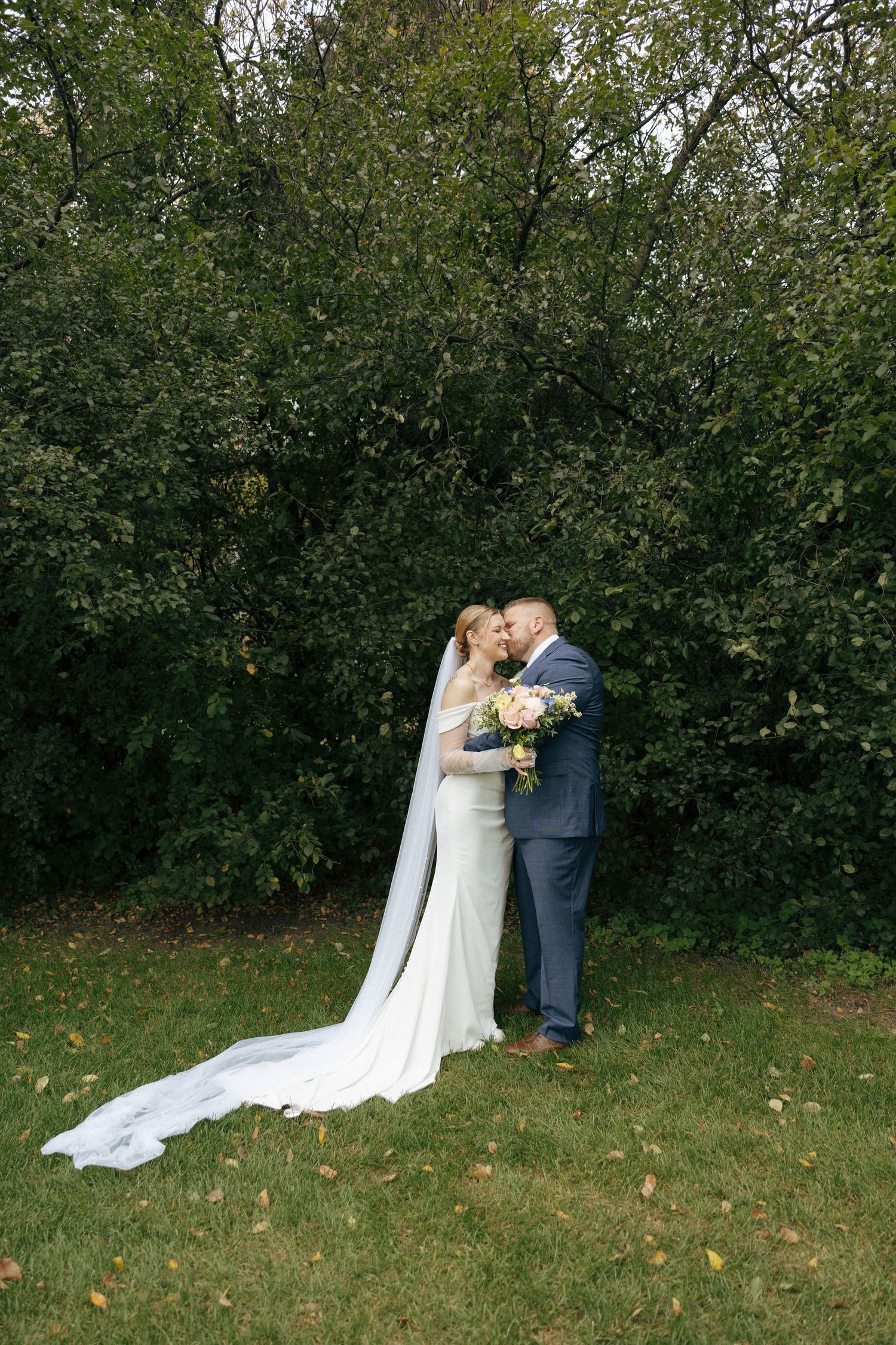 Bride and groom share a kiss outdoors on green grass, with lush trees behind them, as her white veil trails elegantly.