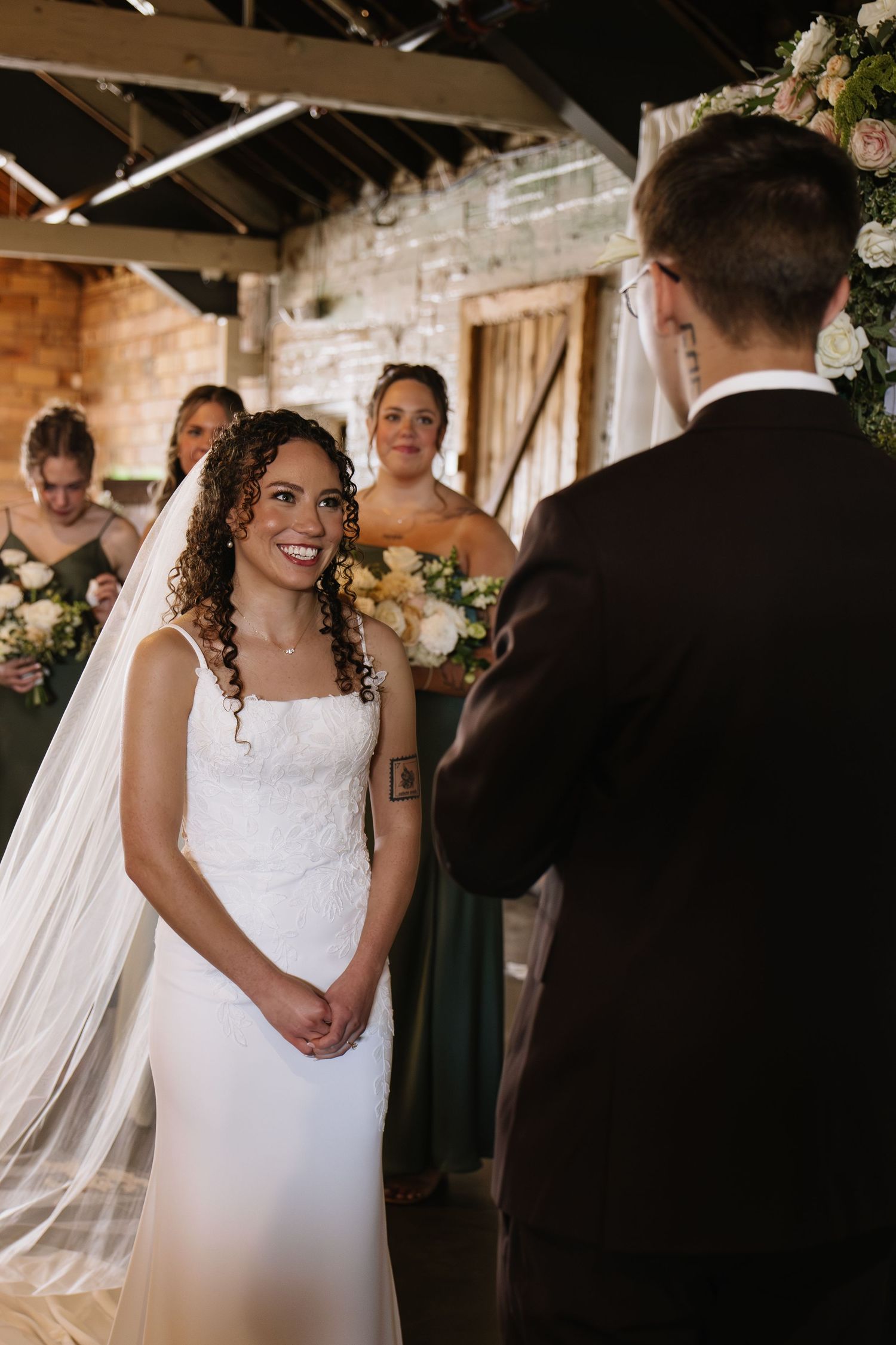 Smiling bride in white gown and veil exchanges vows with groom at rustic indoor wedding ceremony.