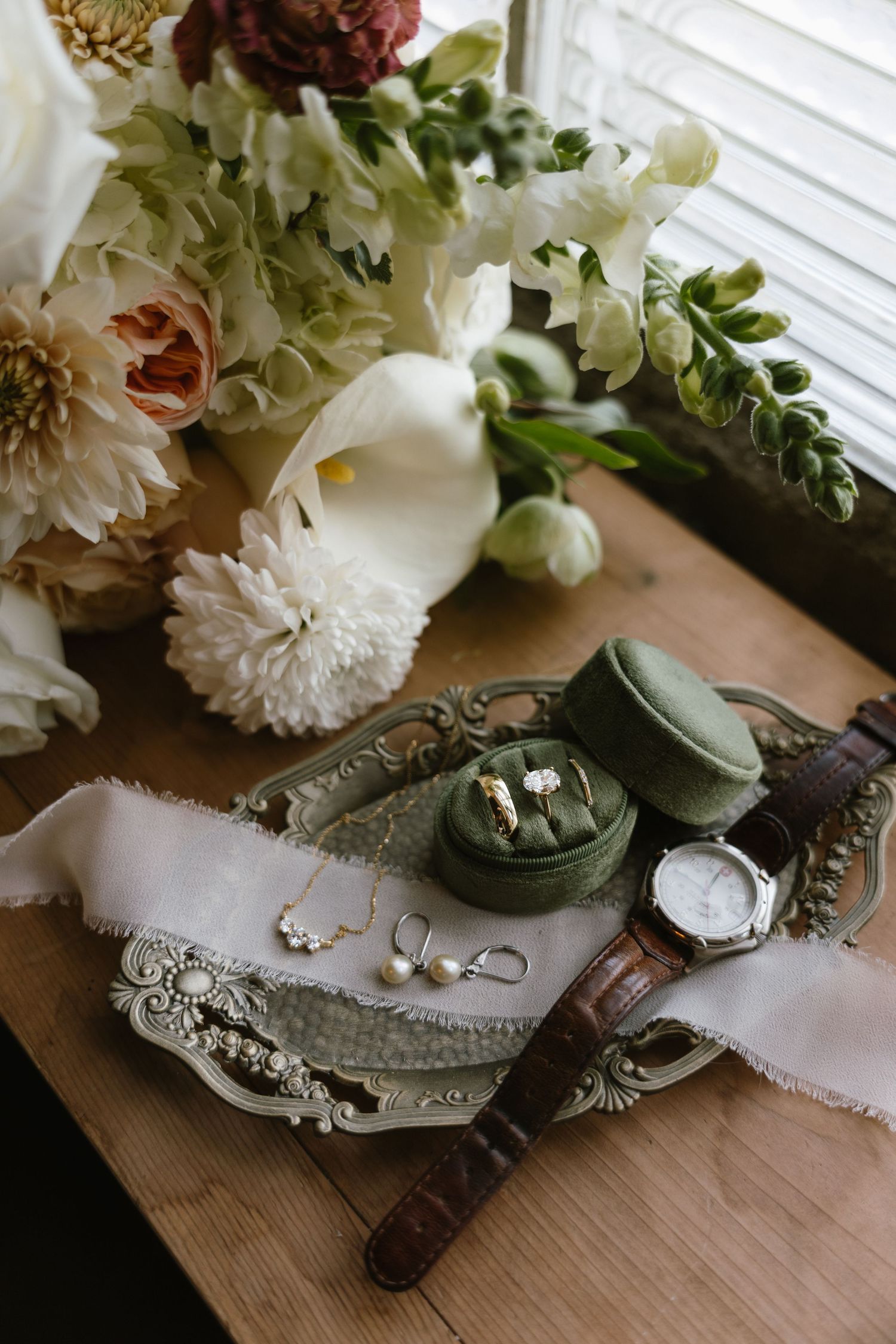 Elegant wedding flat lay with green velvet ring box, pearl earrings, gold necklace, watch, and floral bouquet on ornate tray.