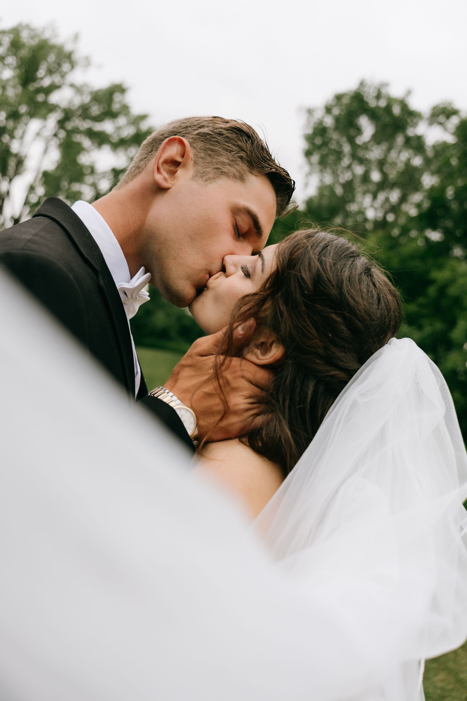 Groom in black suit kissing bride in white veil outdoors with green trees in background.