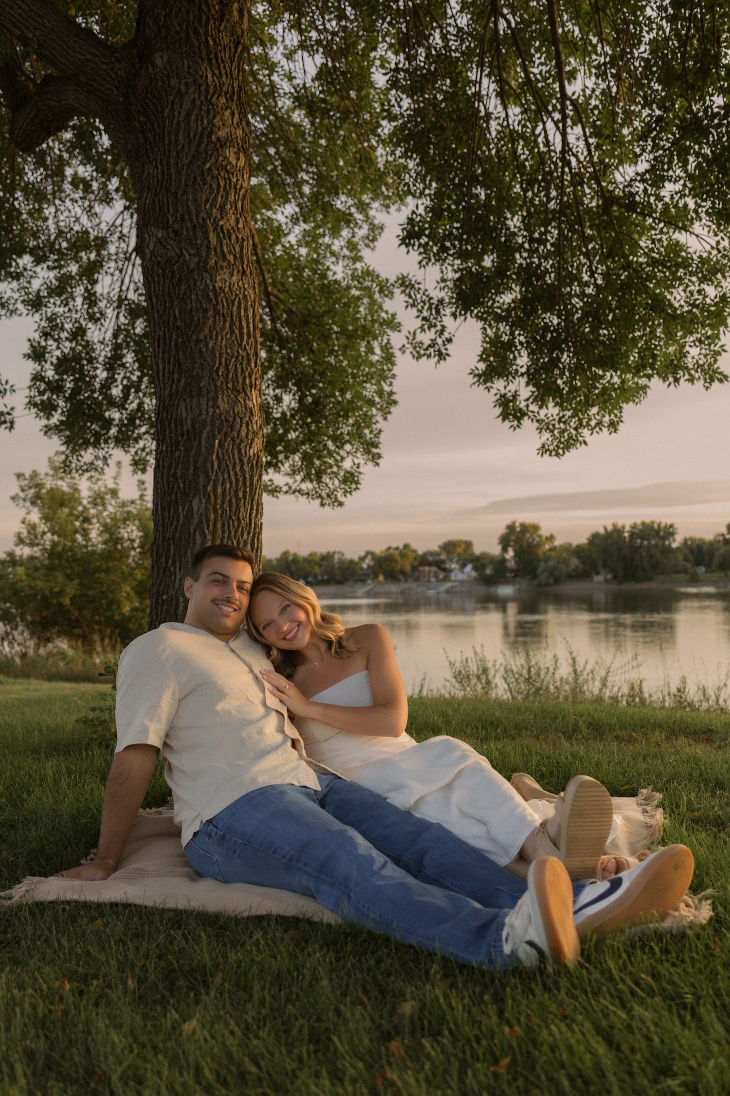A couple relaxes on grass beneath a large tree during golden hour by a lake.