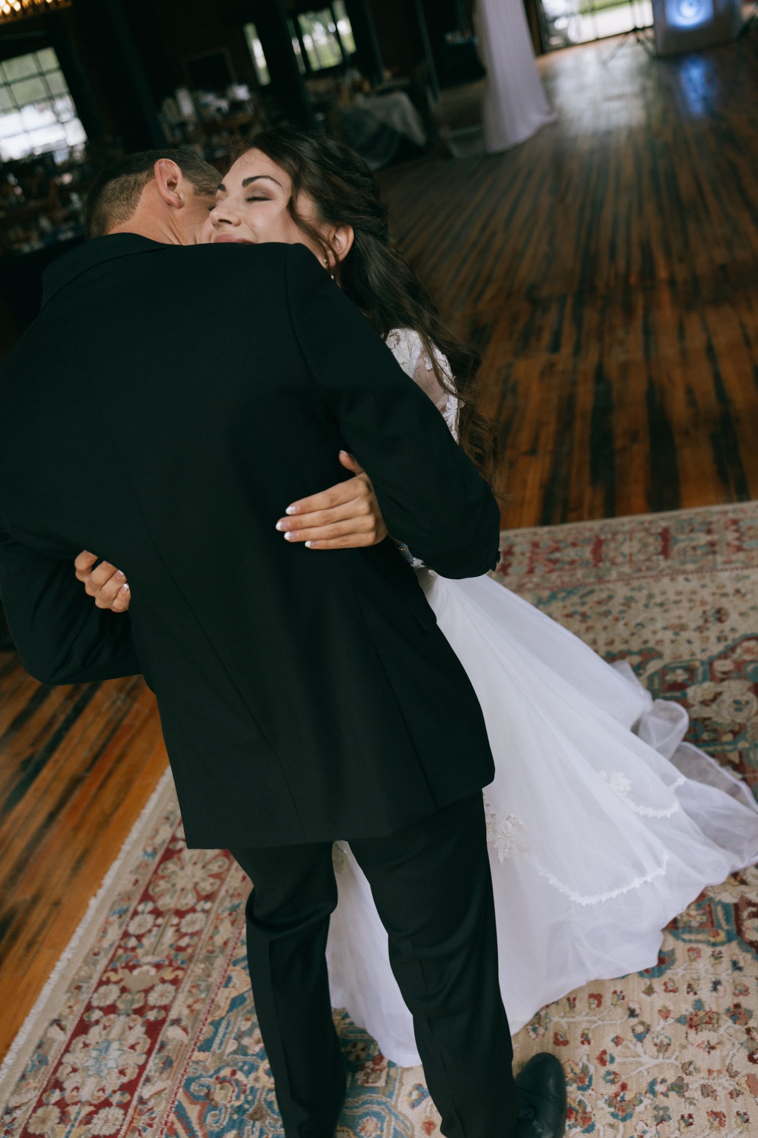 A bride and groom share a joyful first dance embrace on a wooden floor at their wedding reception.