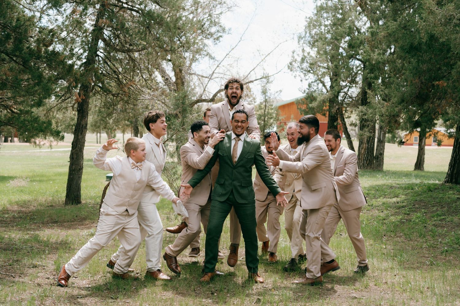 Groom in green suit celebrated by groomsmen in tan suits outdoors among trees at a wedding.