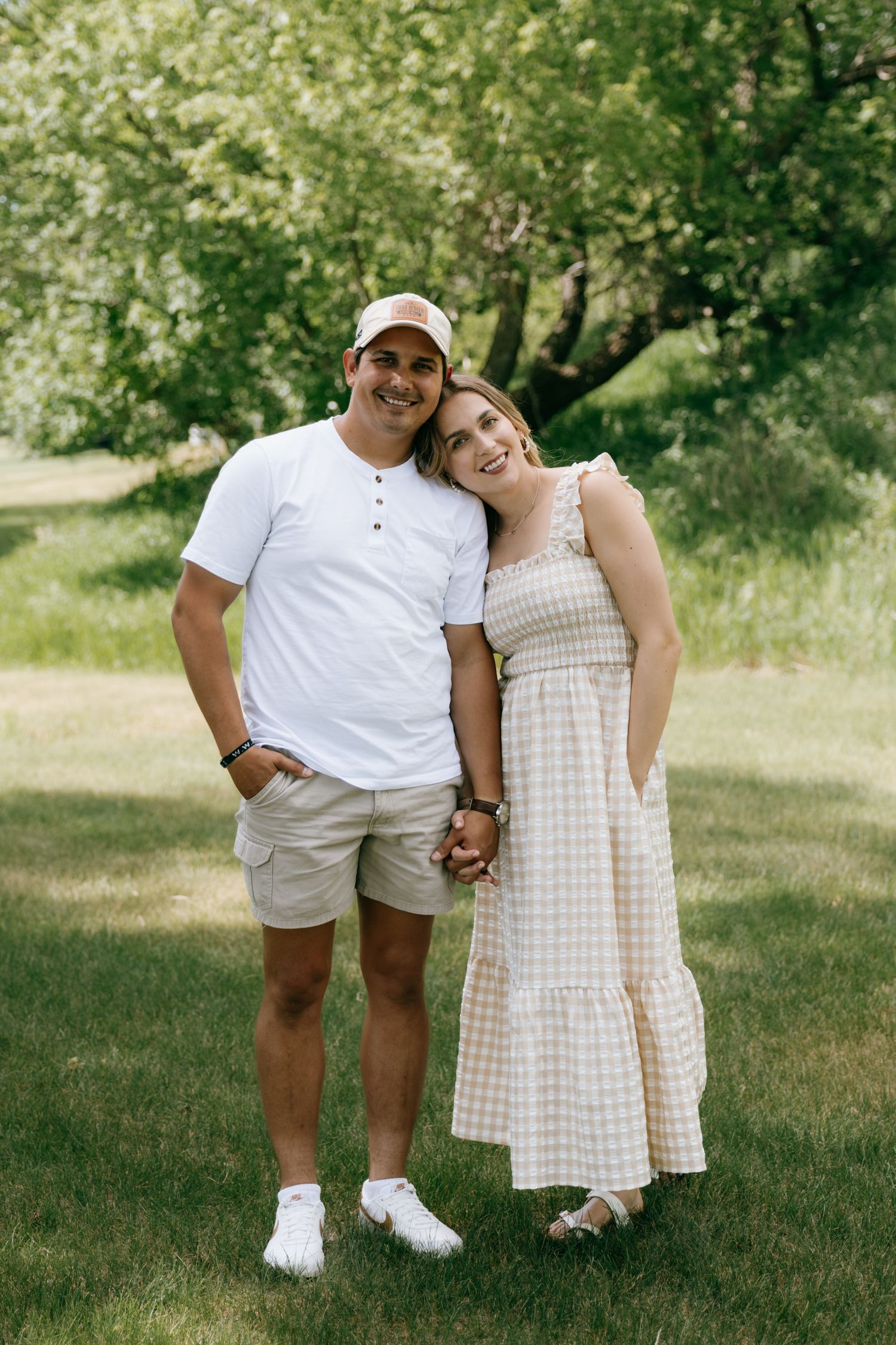 A smiling couple holds hands outdoors on green grass, wearing casual summer outfits, with lush trees in the background.