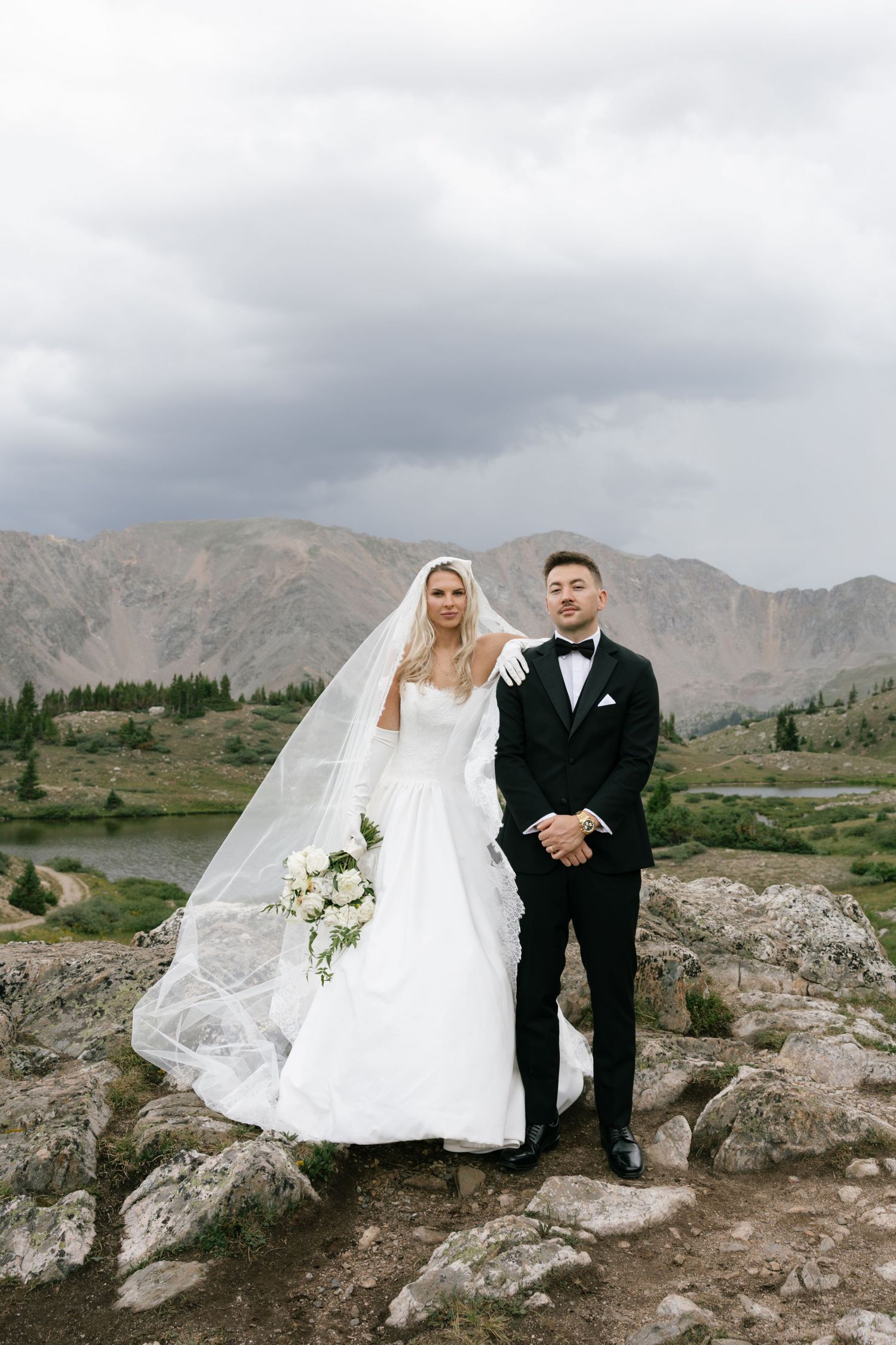 Bride and groom pose on rocky mountain landscape, she in white gown with veil, he in black tuxedo.