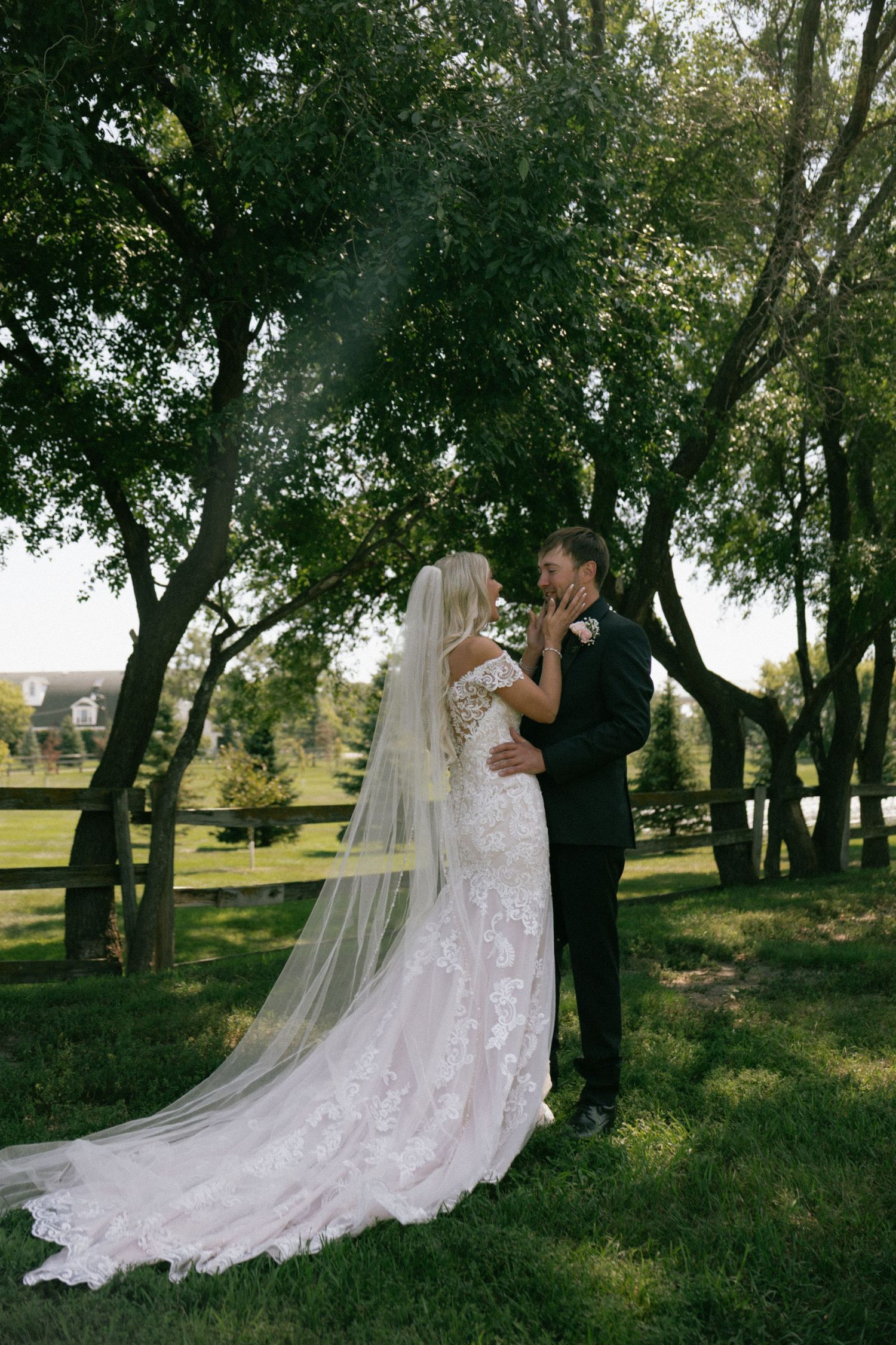 A bride in a lace gown and veil shares a romantic moment with her groom in a suit outdoors.