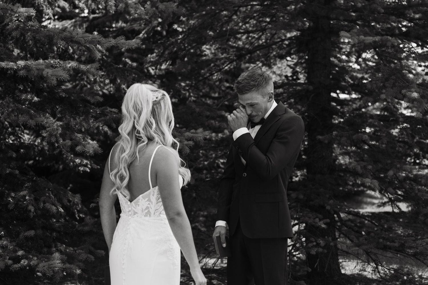 Groom wiping tears during emotional first look with bride in white lace gown among evergreen trees.