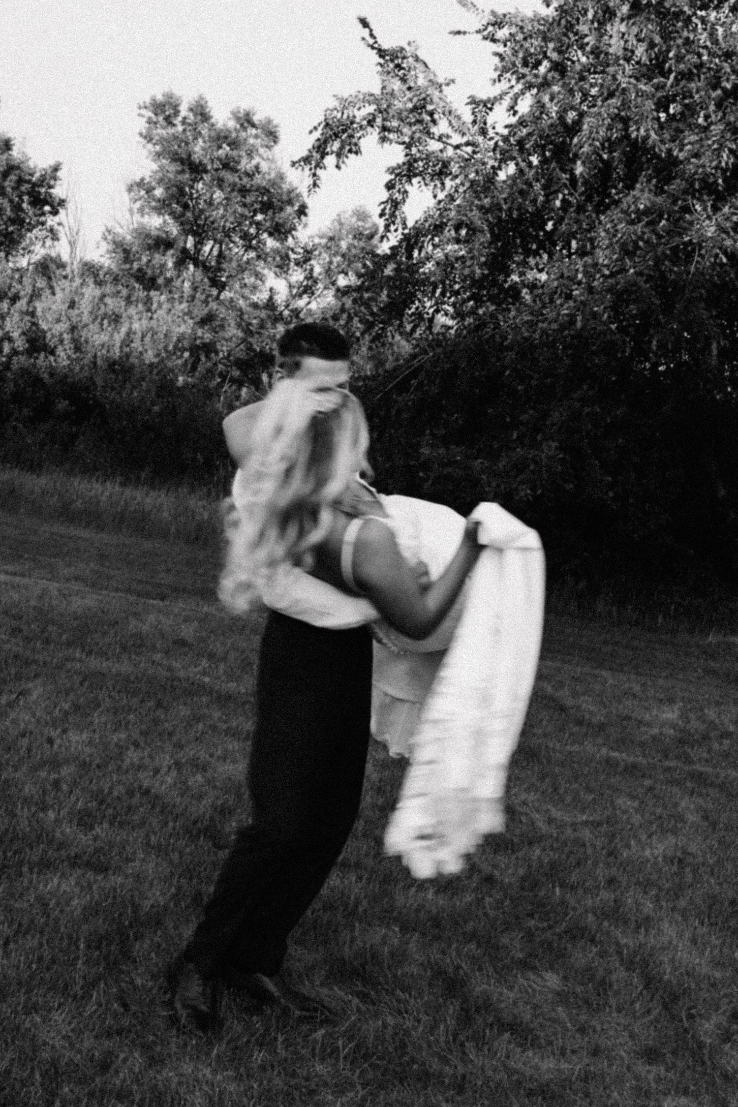 Black and white photo of a couple dancing outdoors, bride in flowing white dress being spun by groom near trees.