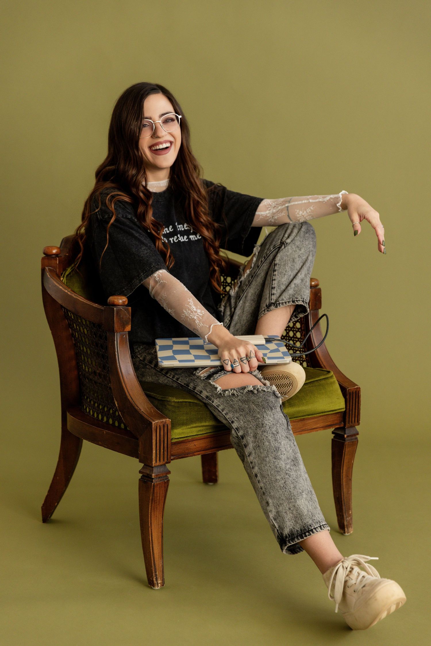 Smiling young woman with tattoos sits in wooden chair, wearing black tee, ripped jeans, glasses, holding checkered clutch.