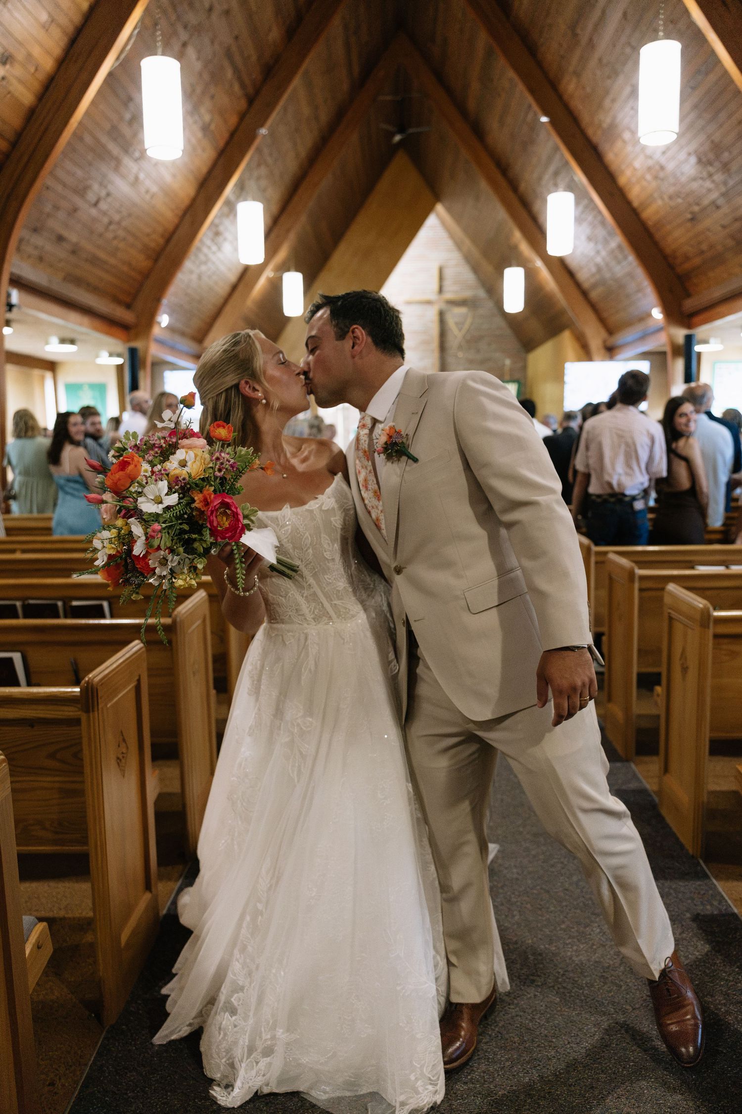 Bride and groom share a kiss in church aisle, bride holding colorful bouquet, groom in beige suit, wooden chapel.