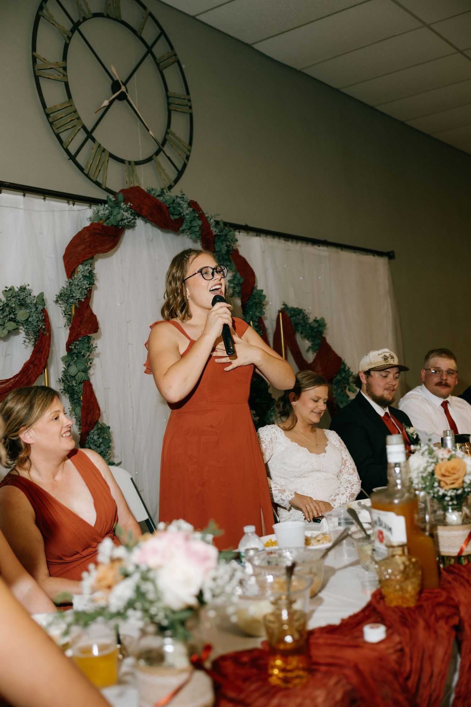A bridesmaid in a rust dress gives a toast at a wedding reception with greenery and red decor backdrop.