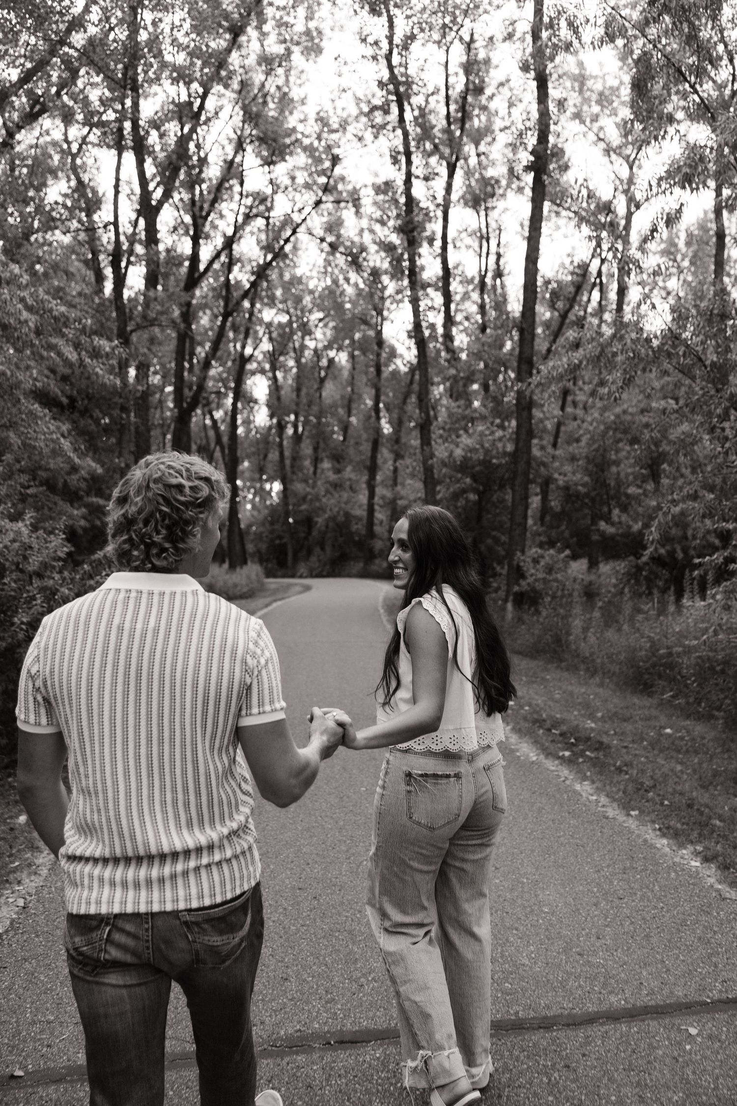 Black and white photo of a couple holding hands while walking on a park path surrounded by tall trees.