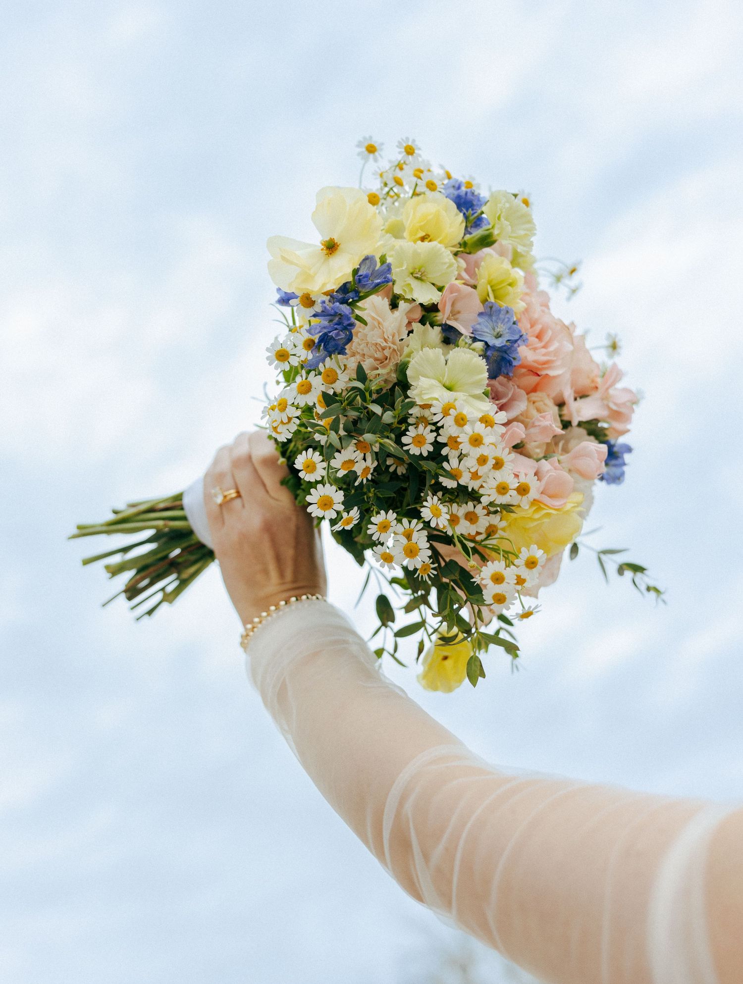 A hand holding a vibrant spring wedding bouquet with daisies, blue, pink, and yellow flowers against a cloudy sky.