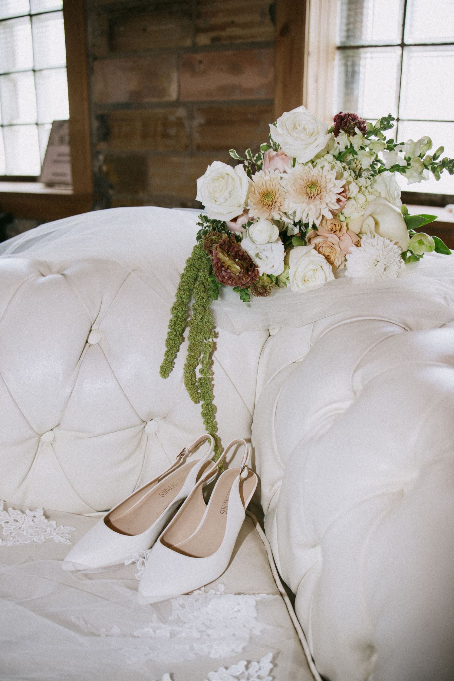 Elegant bridal heels and white floral bouquet with greenery resting on a tufted ivory velvet sofa.