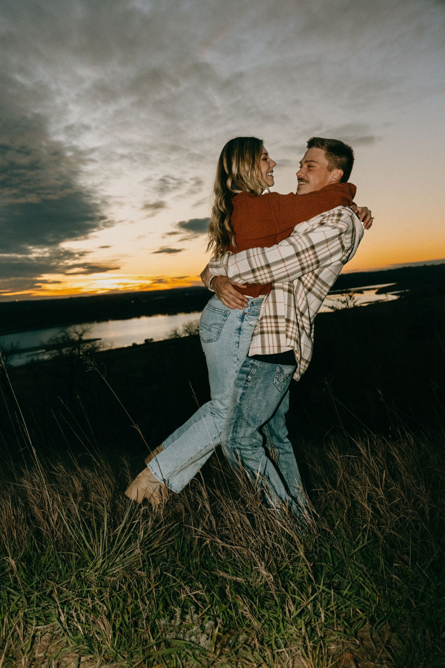 Couple in plaid and denim share playful embrace in tall grass during golden hour.