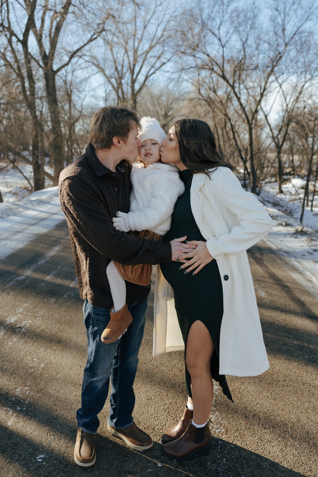 photos of mom and dad kissing cheeks of daughter in snowy trees in Bismarck