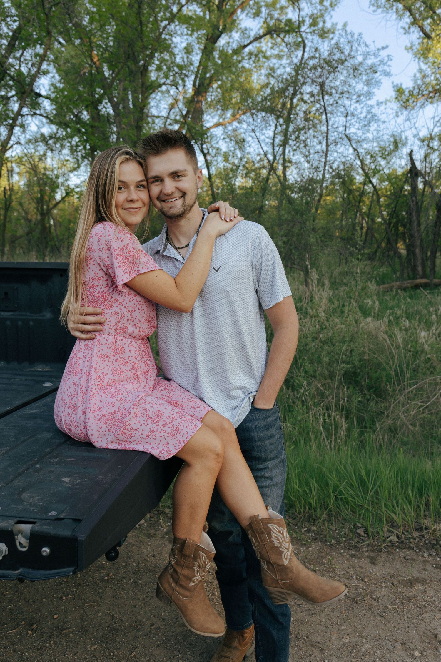 Couple poses casually on black truck bed in outdoor nature setting.