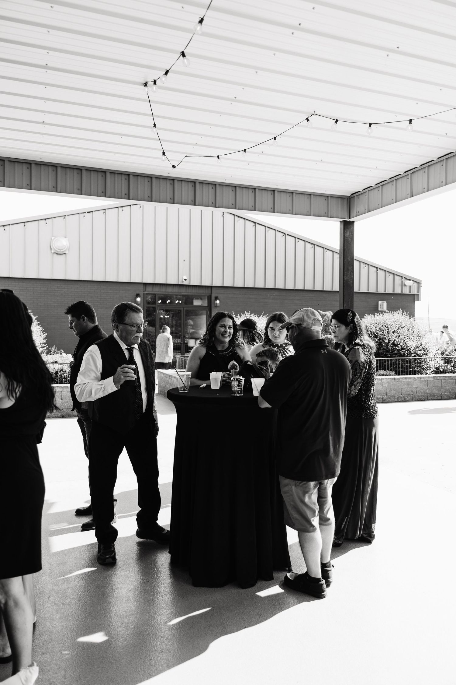 Black and white photo of elegantly dressed guests mingling outdoors at a formal event under string lights.