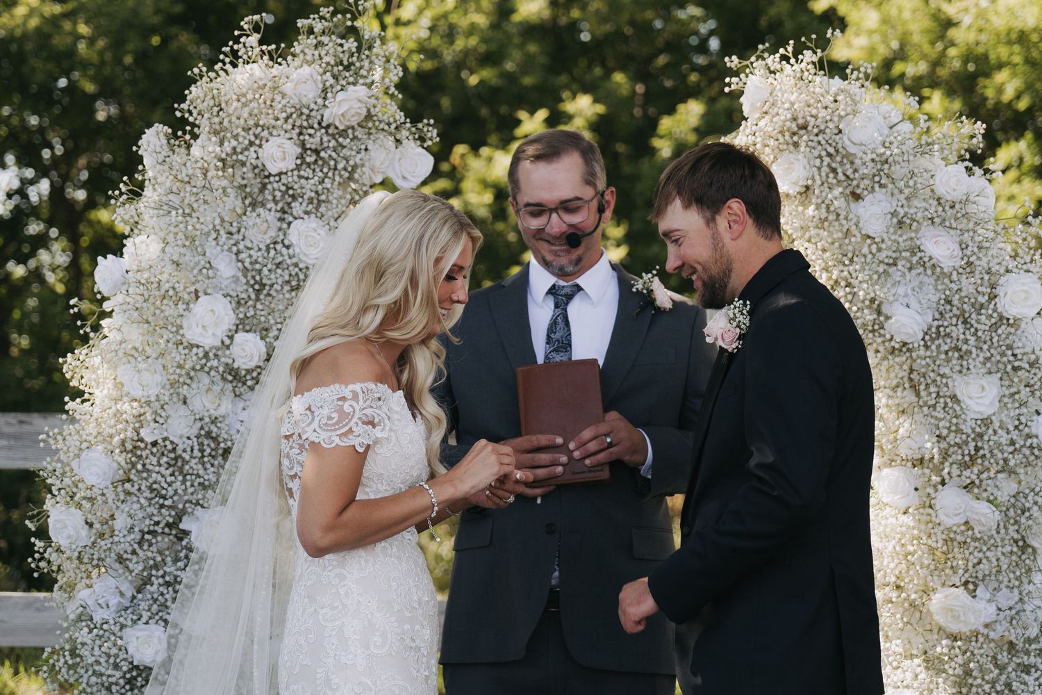 Bride and groom exchange rings during outdoor wedding ceremony beneath floral arch with white roses and baby's breath.