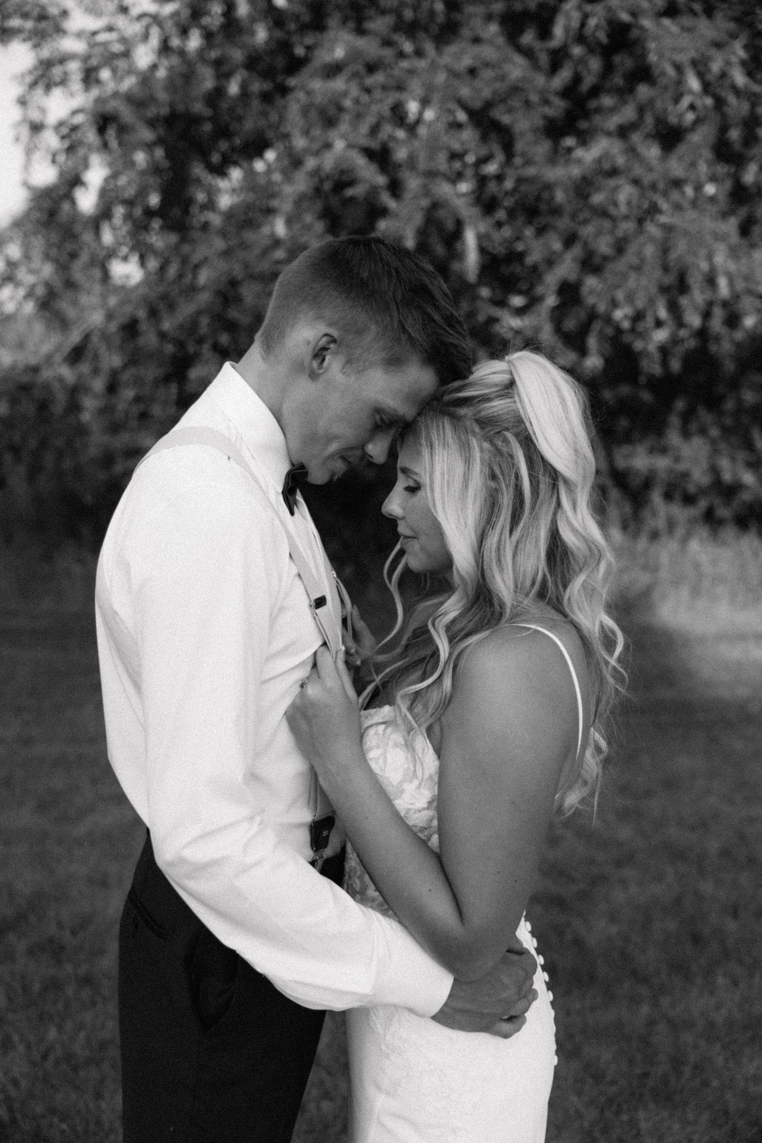Black and white wedding photo of bride and groom embracing outdoors, sharing an intimate moment among lush trees.