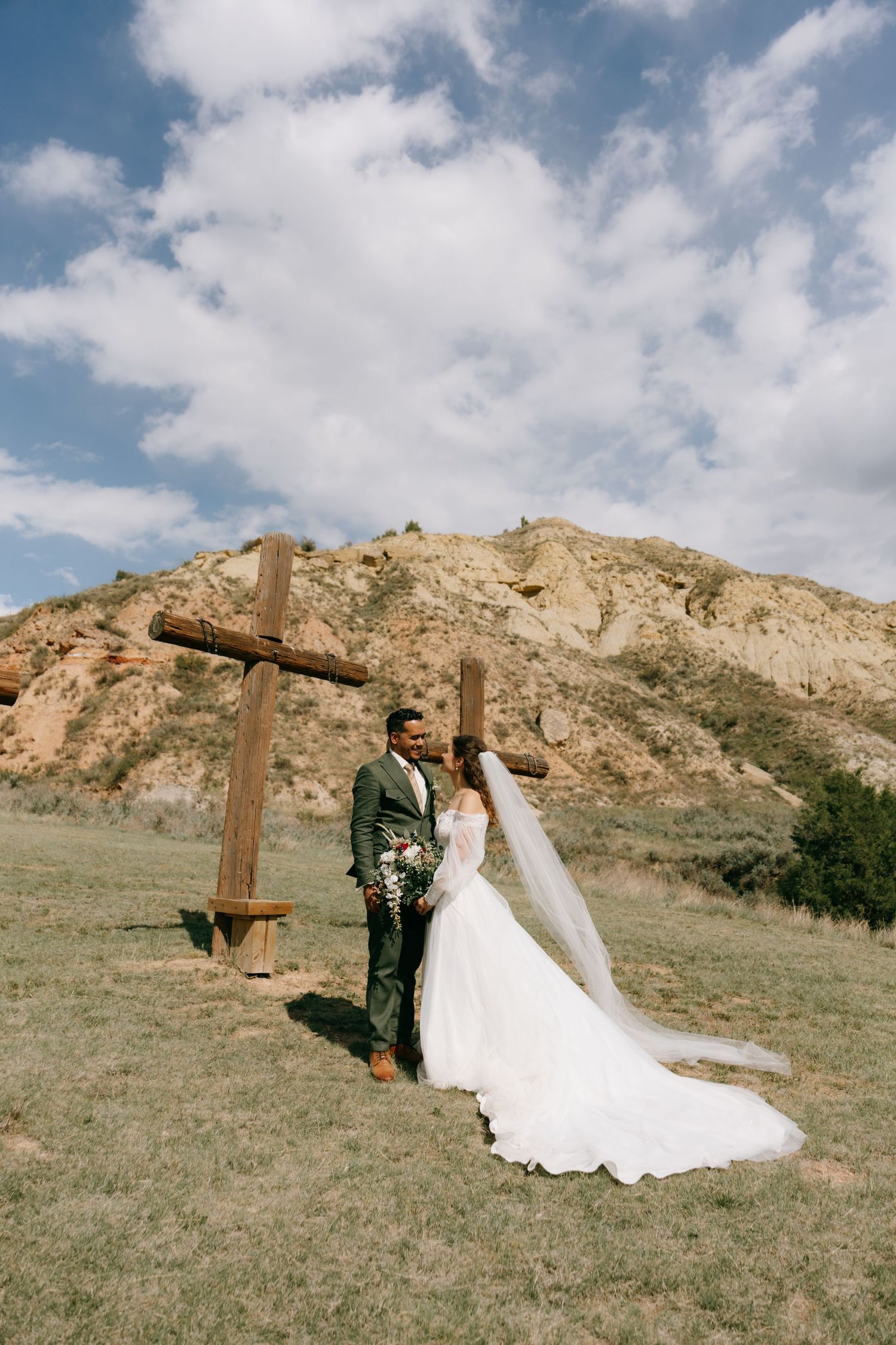 Bride and groom pose before rustic wooden cross on scenic hillside during outdoor wedding ceremony.