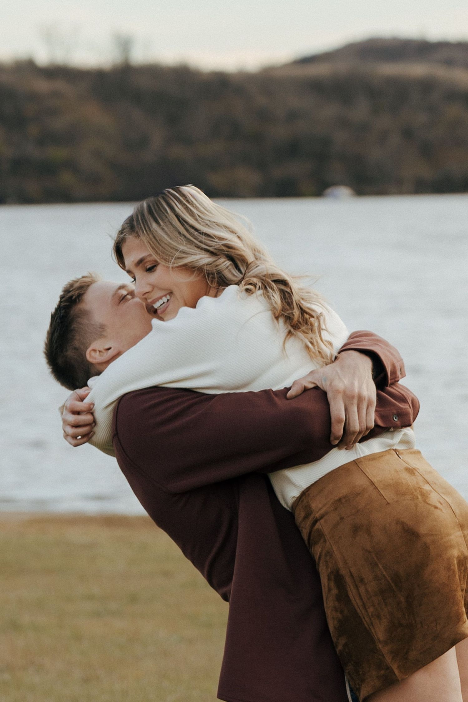 A couple shares an intimate embrace in a field near a lake during golden hour.