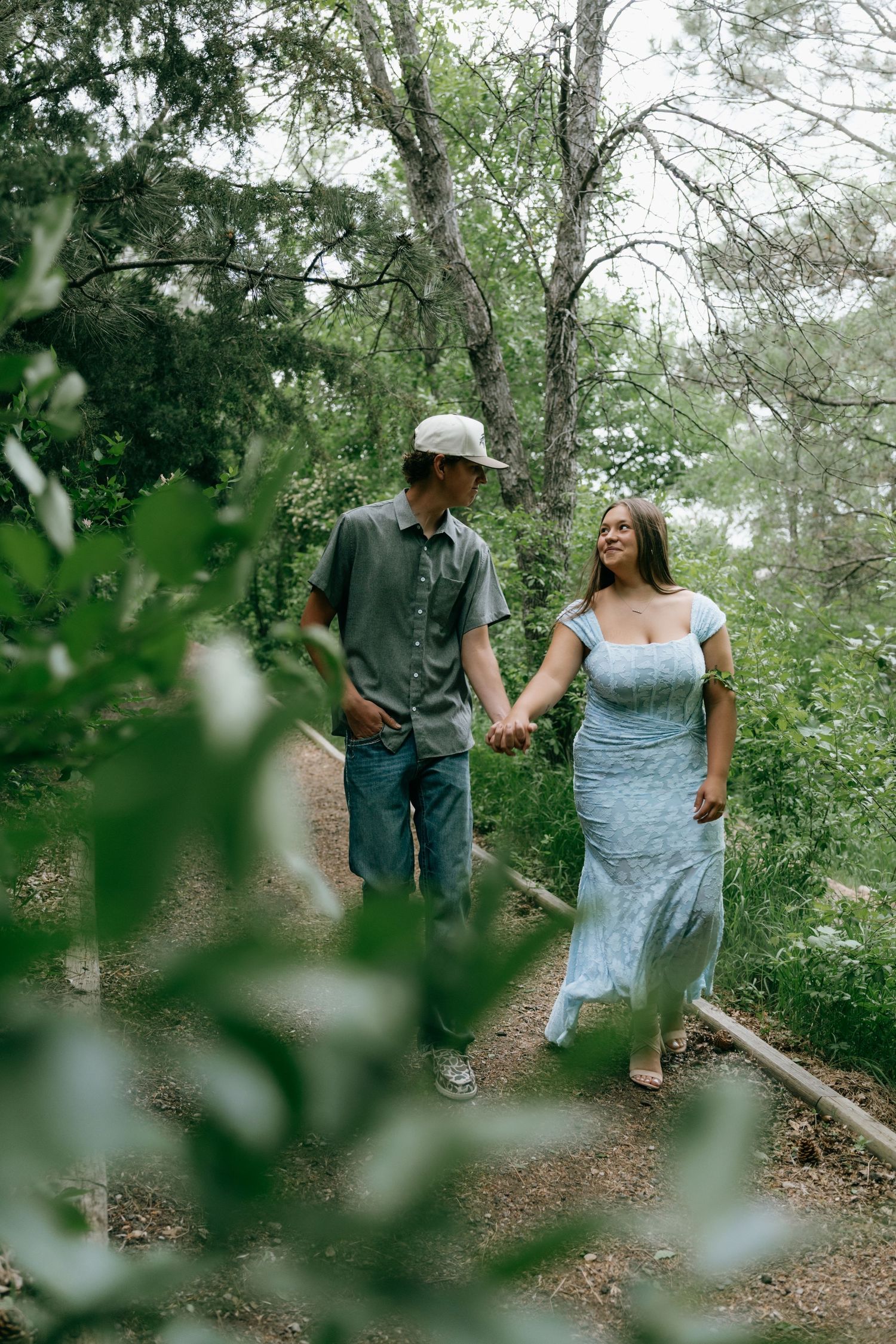 A couple holds hands and smiles while walking on a forest trail, woman in blue dress and man in hat.
