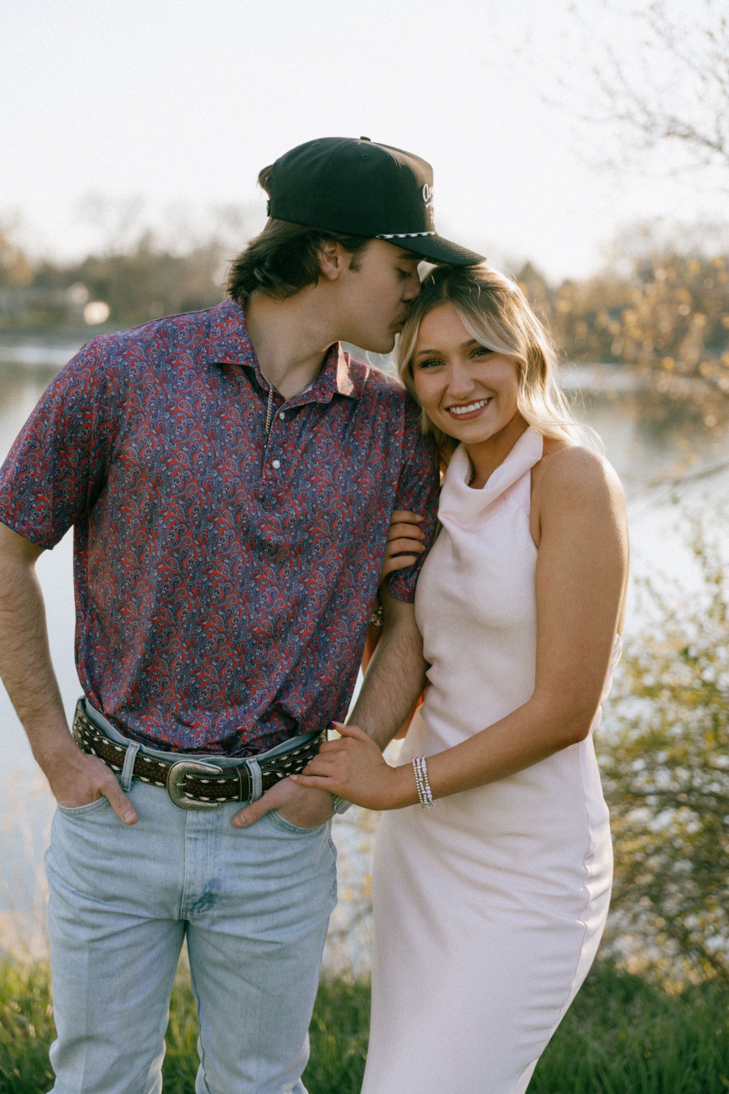 Couple posing by a lake at sunset, man kissing woman's cheek as she smiles in a white dress.