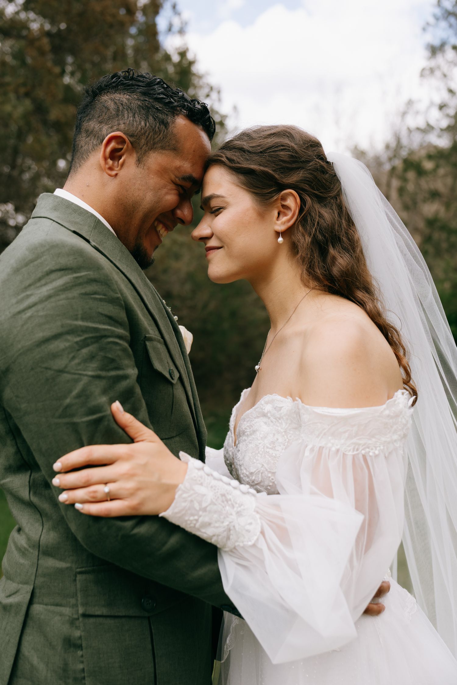 A bride and groom share a romantic forehead touch, smiling outdoors, she in a white lace gown and he in a green suit.