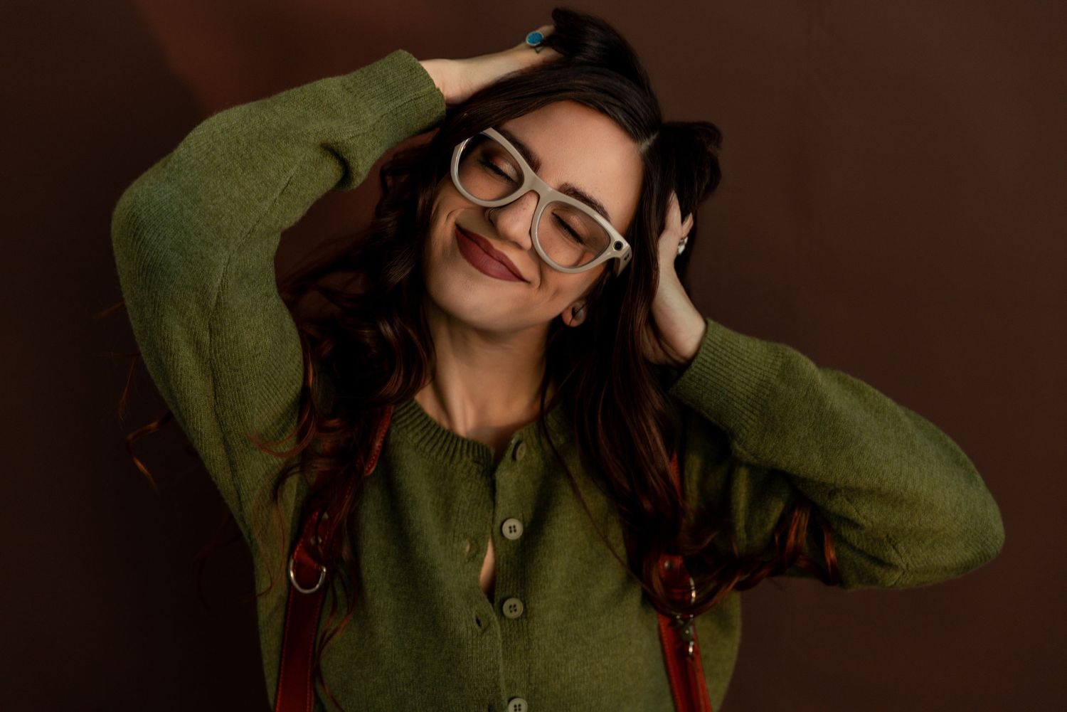 A smiling woman with glasses and long brown hair wearing a green cardigan poses with hands on her head against a dark background.