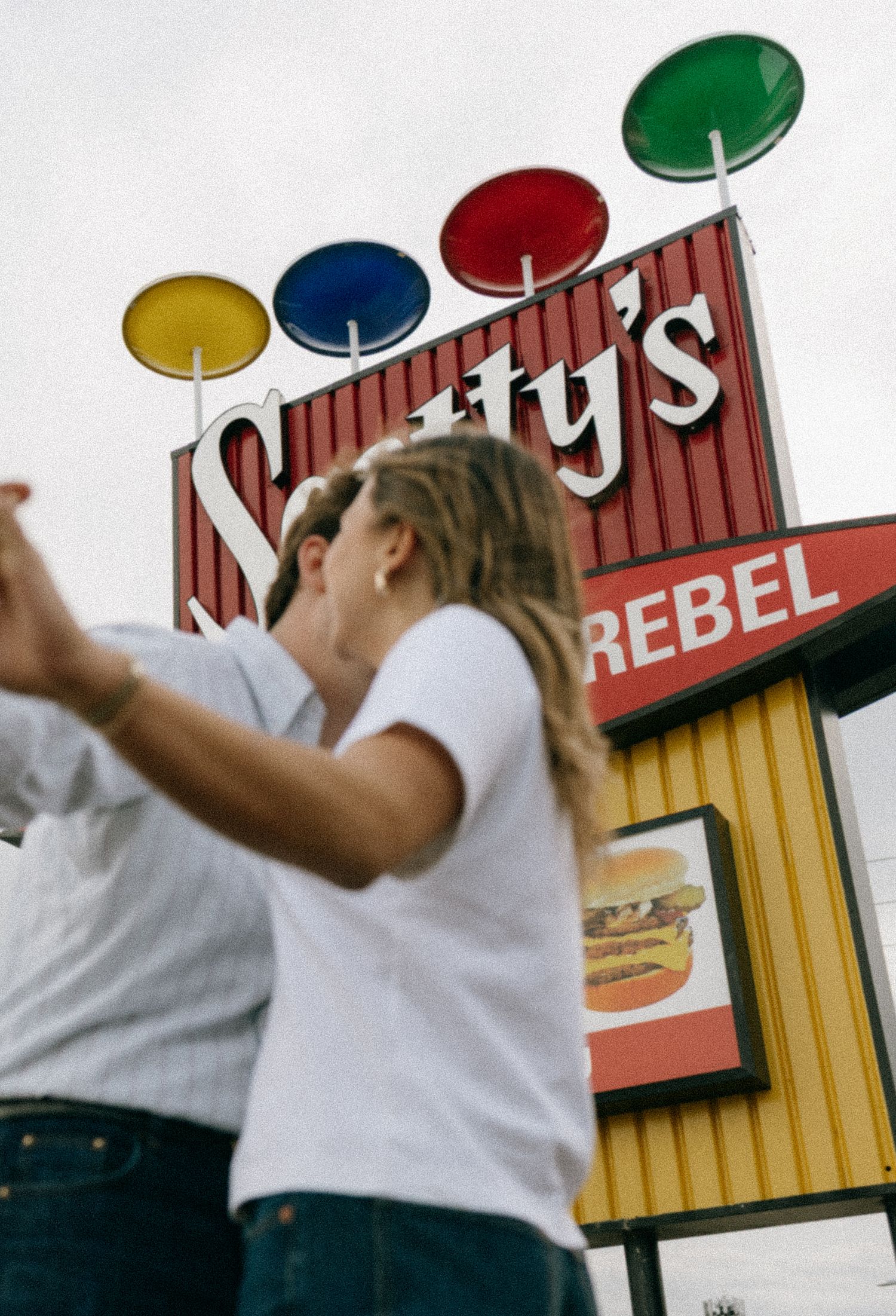 Two people stand in front of a colorful Scotty's fast food restaurant sign with large lollipop decorations.