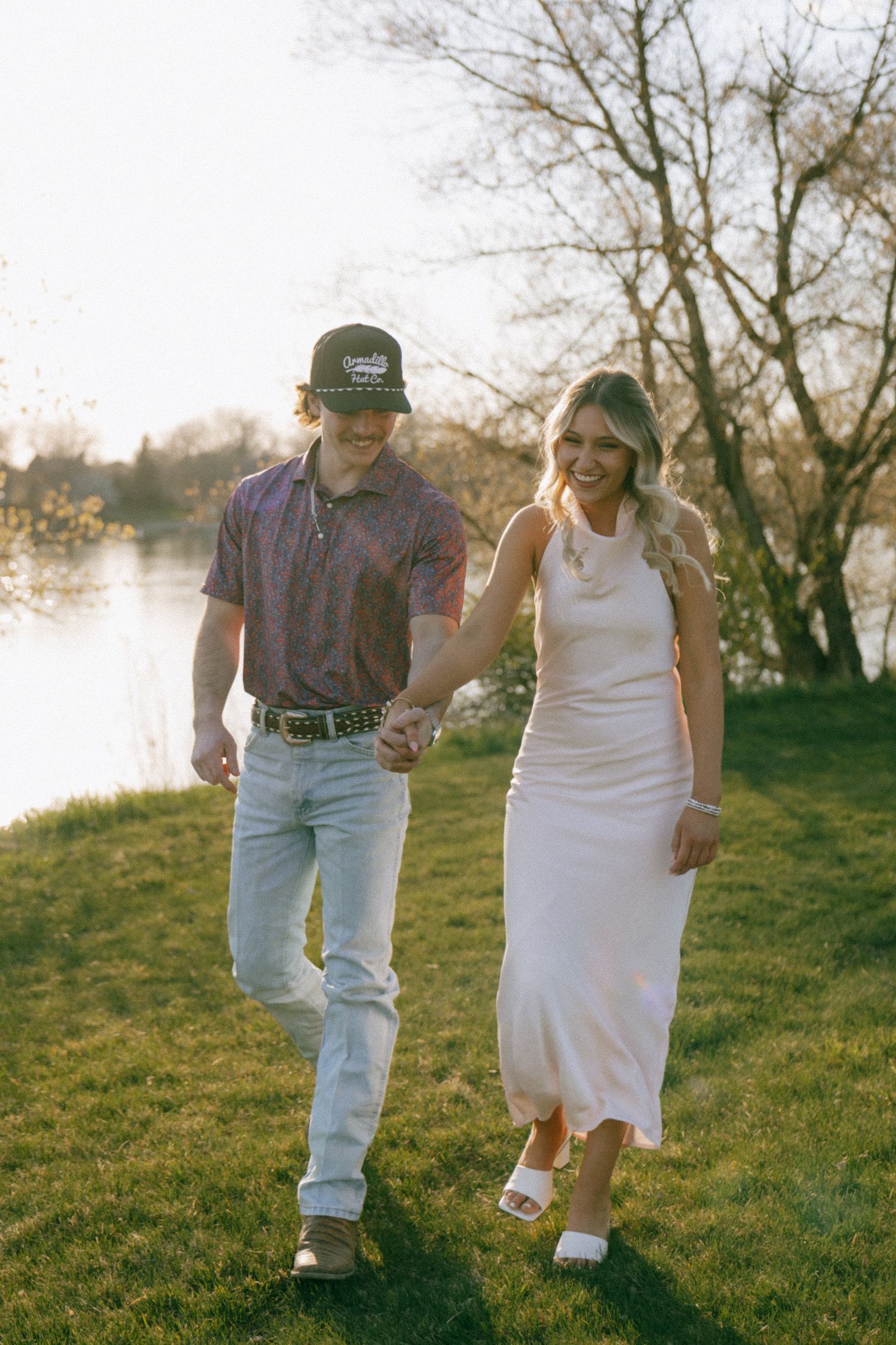 Couple holding hands walking on grass near a lake, woman in white dress and man in purple shirt and cap.