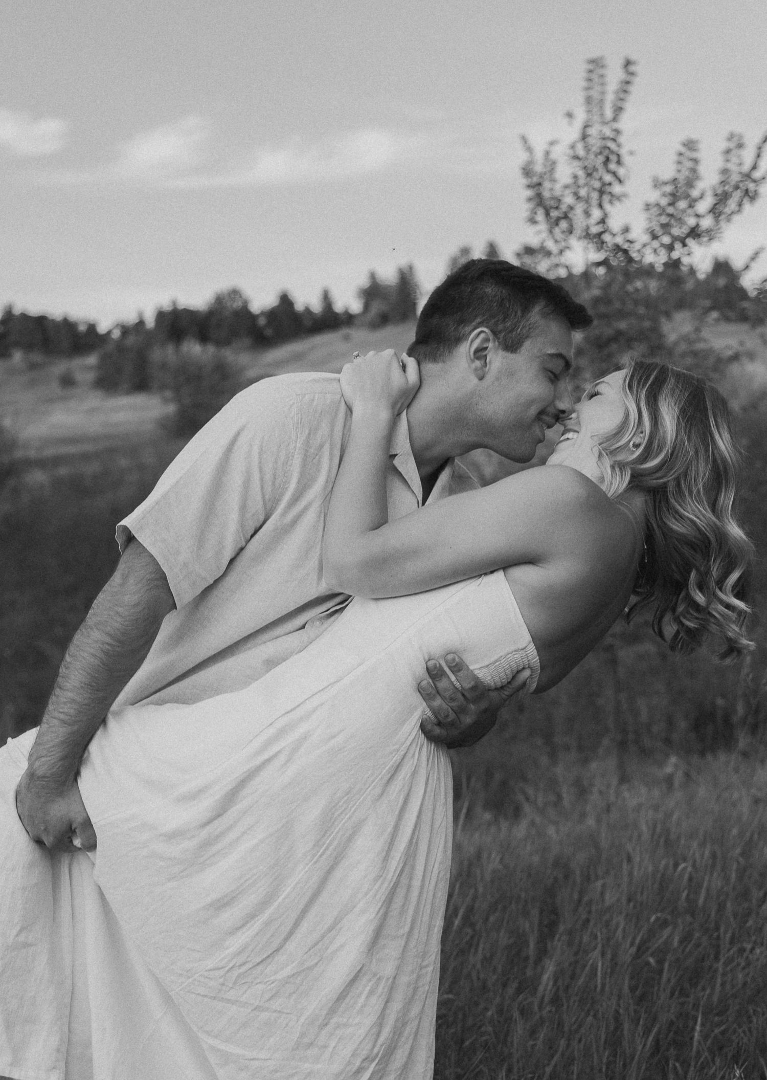 A romantic black and white portrait of a couple sharing an intimate dip kiss in a rustic outdoor field setting.