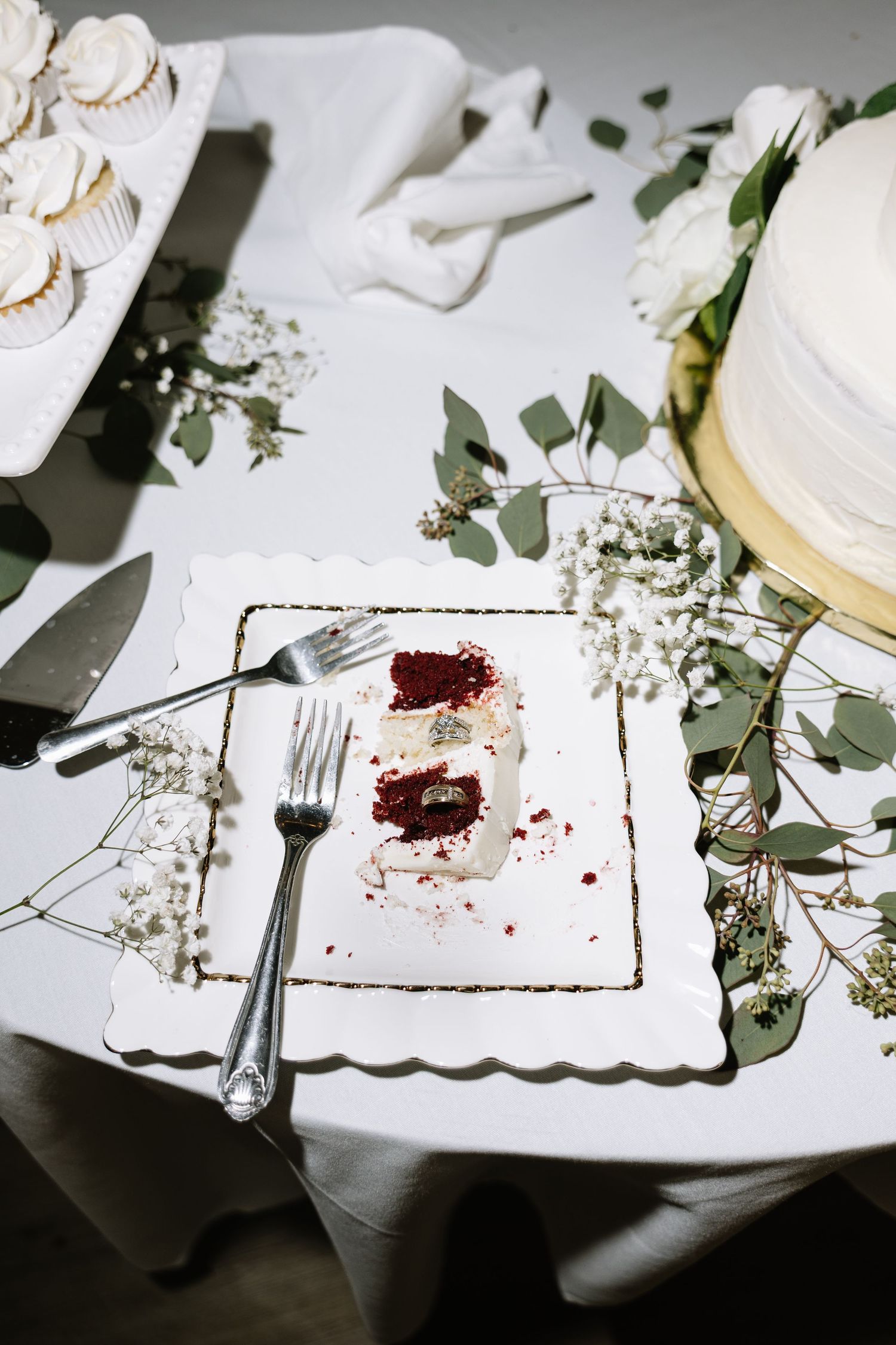 Elegant wedding cake slice on white plate with forks, surrounded by eucalyptus and baby's breath floral decor.