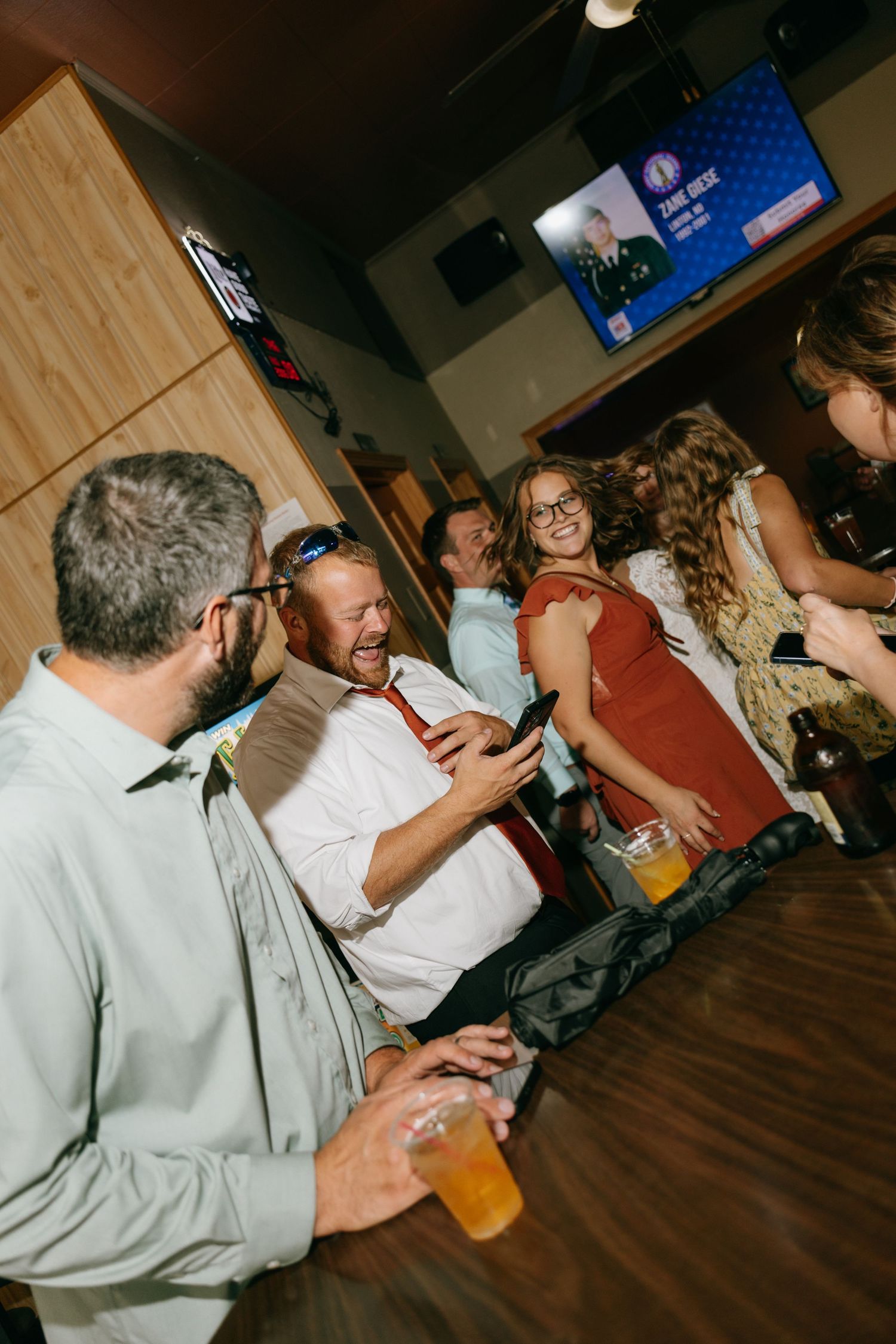 Group of friends laughing and socializing at a bar, with drinks in hand and a TV screen visible in the background.