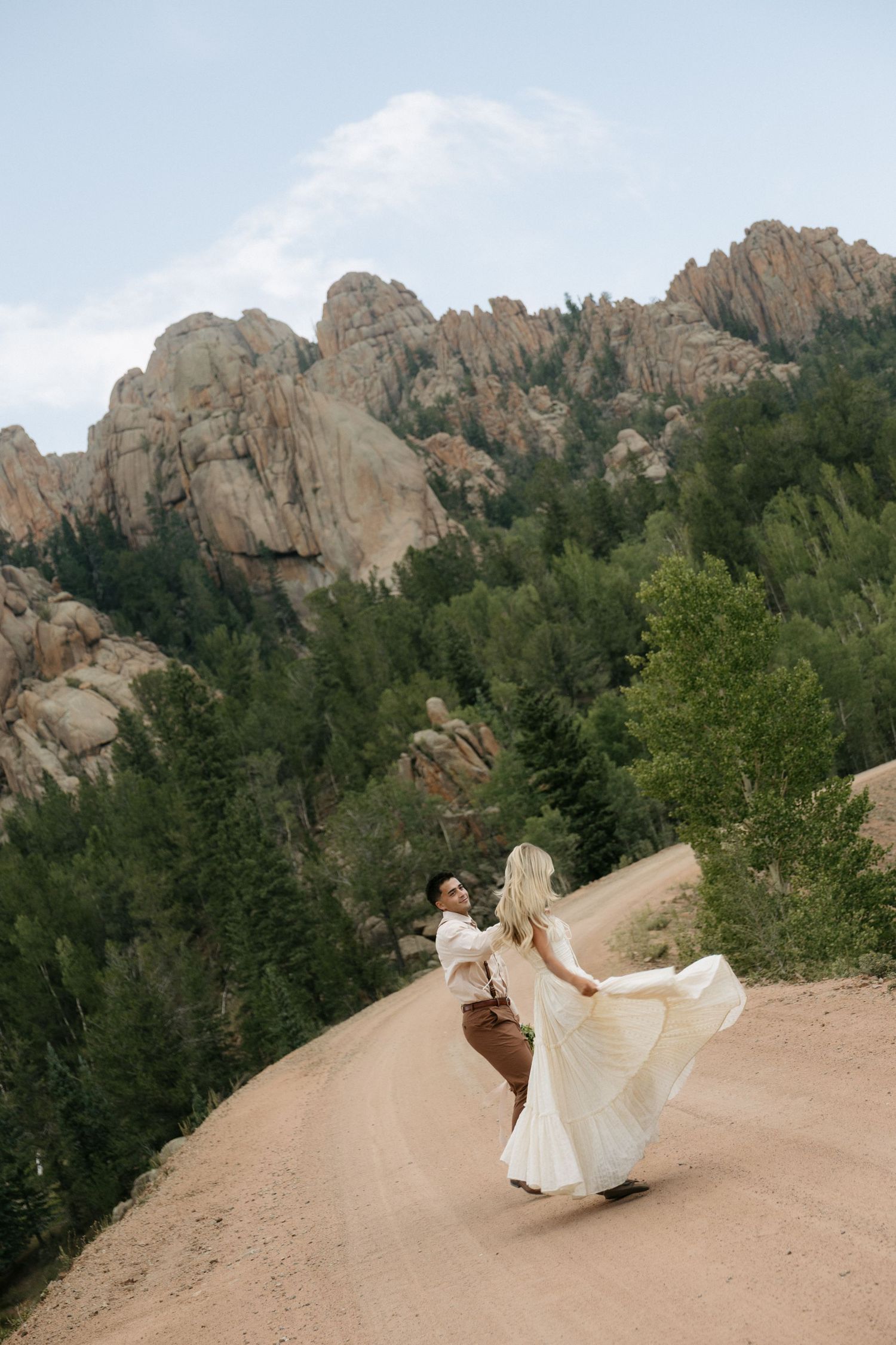 Bride and groom dancing on a dirt road surrounded by rocky mountains and pine trees on their wedding day.