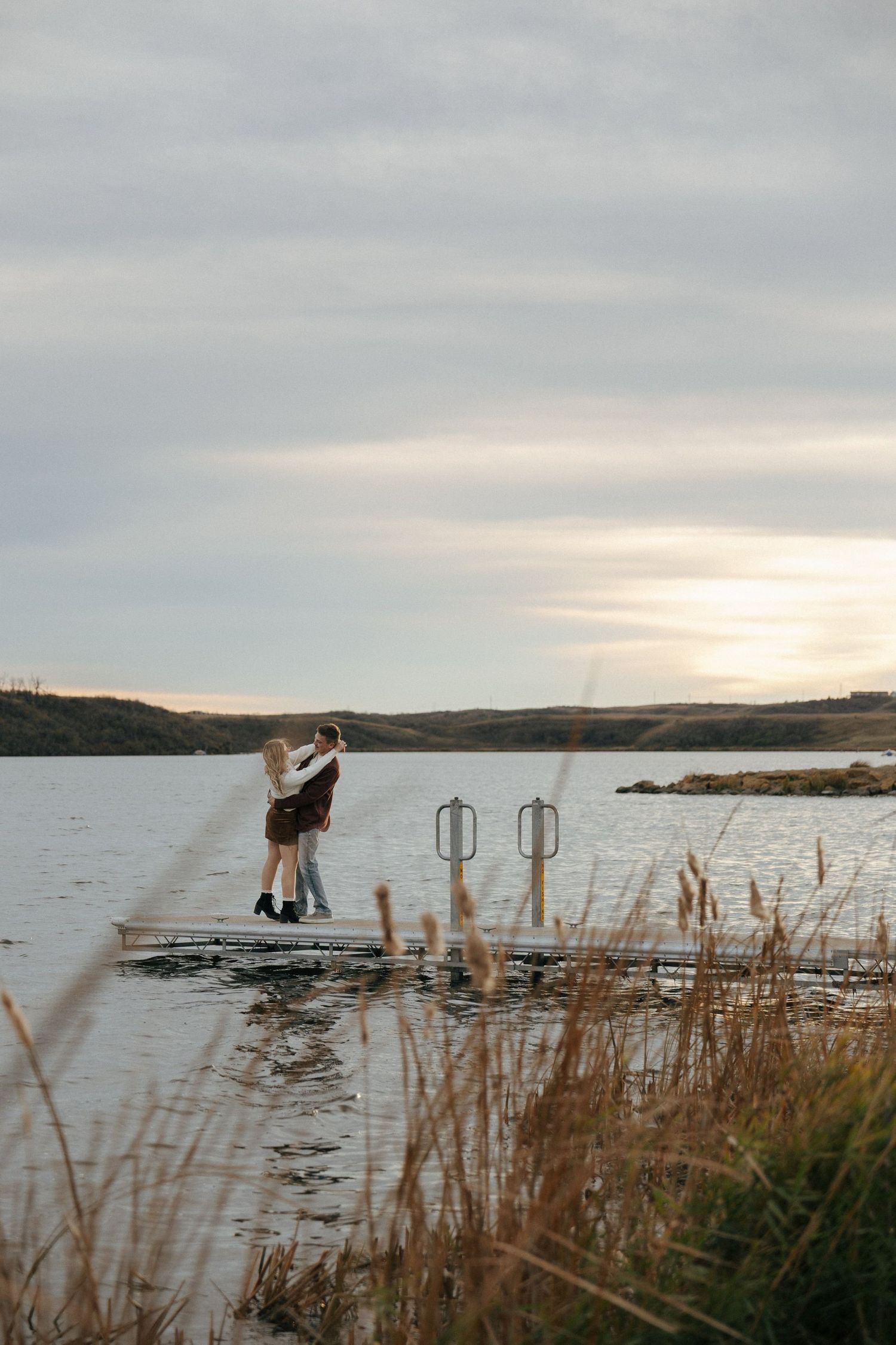 A romantic lakeside scene with a couple embracing near the water's edge at dusk.