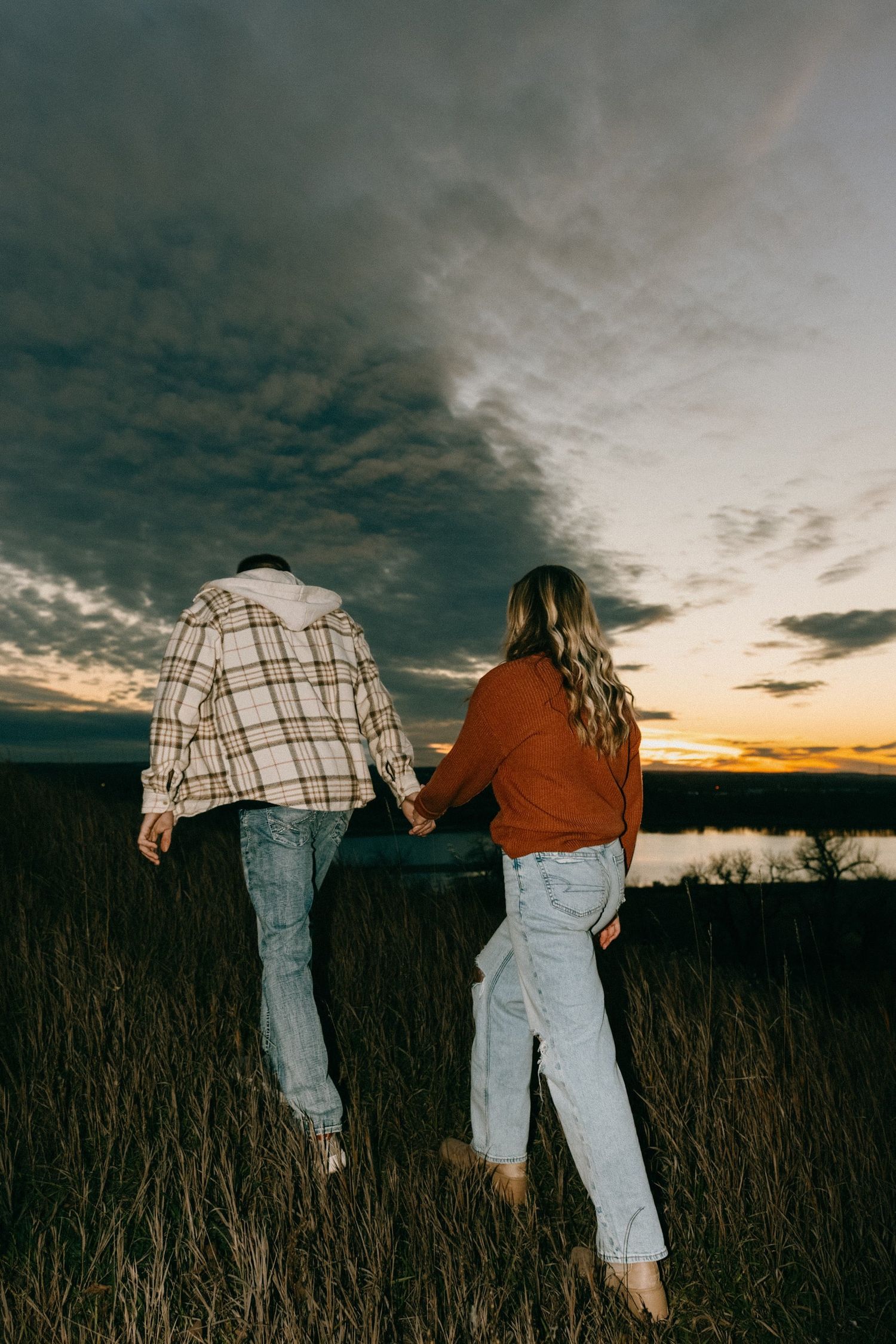 Couple holding hands while walking through tall grass with lake and sunset view.