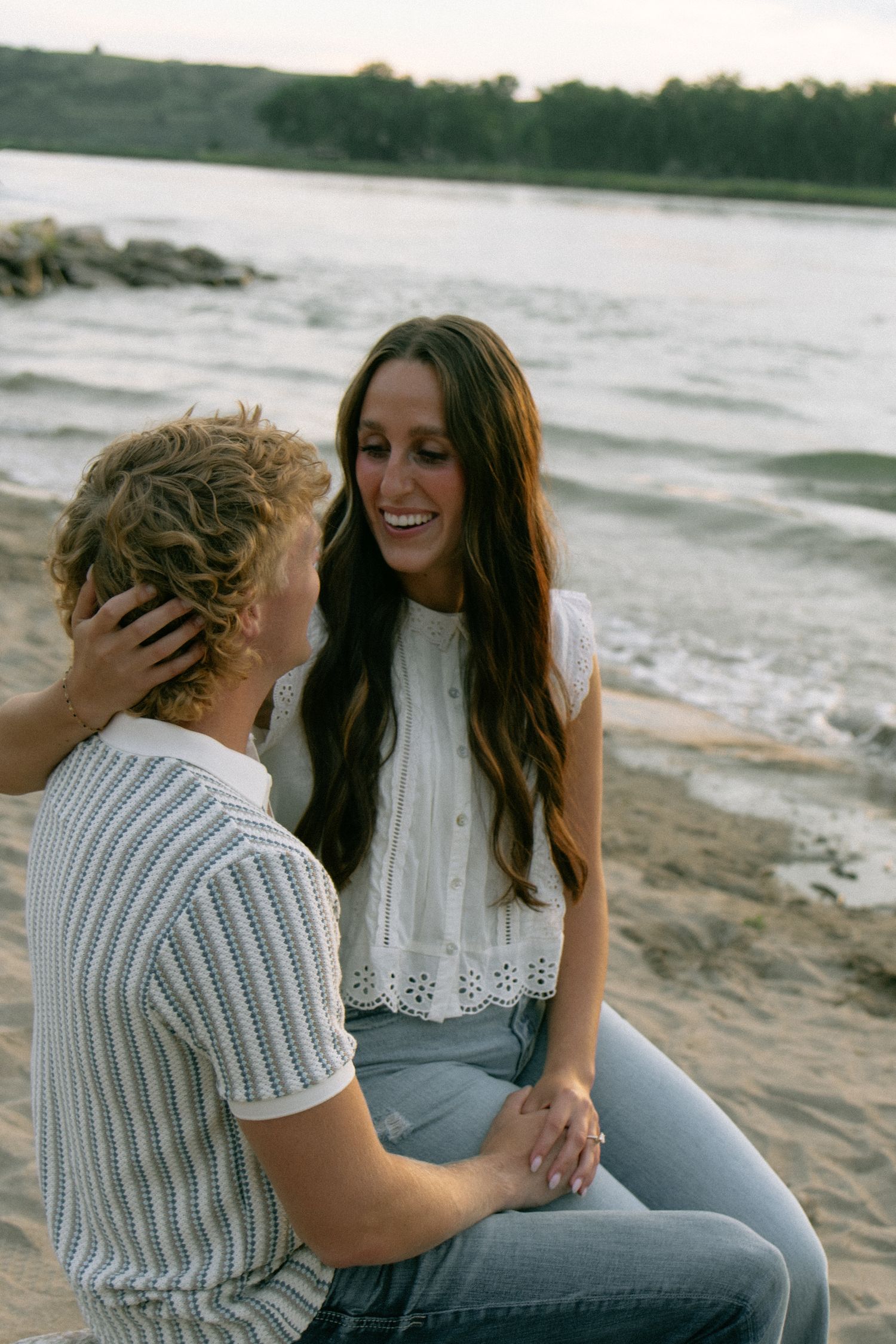 A smiling couple sits on a sandy beach at dusk, holding hands and gazing at each other affectionately.
