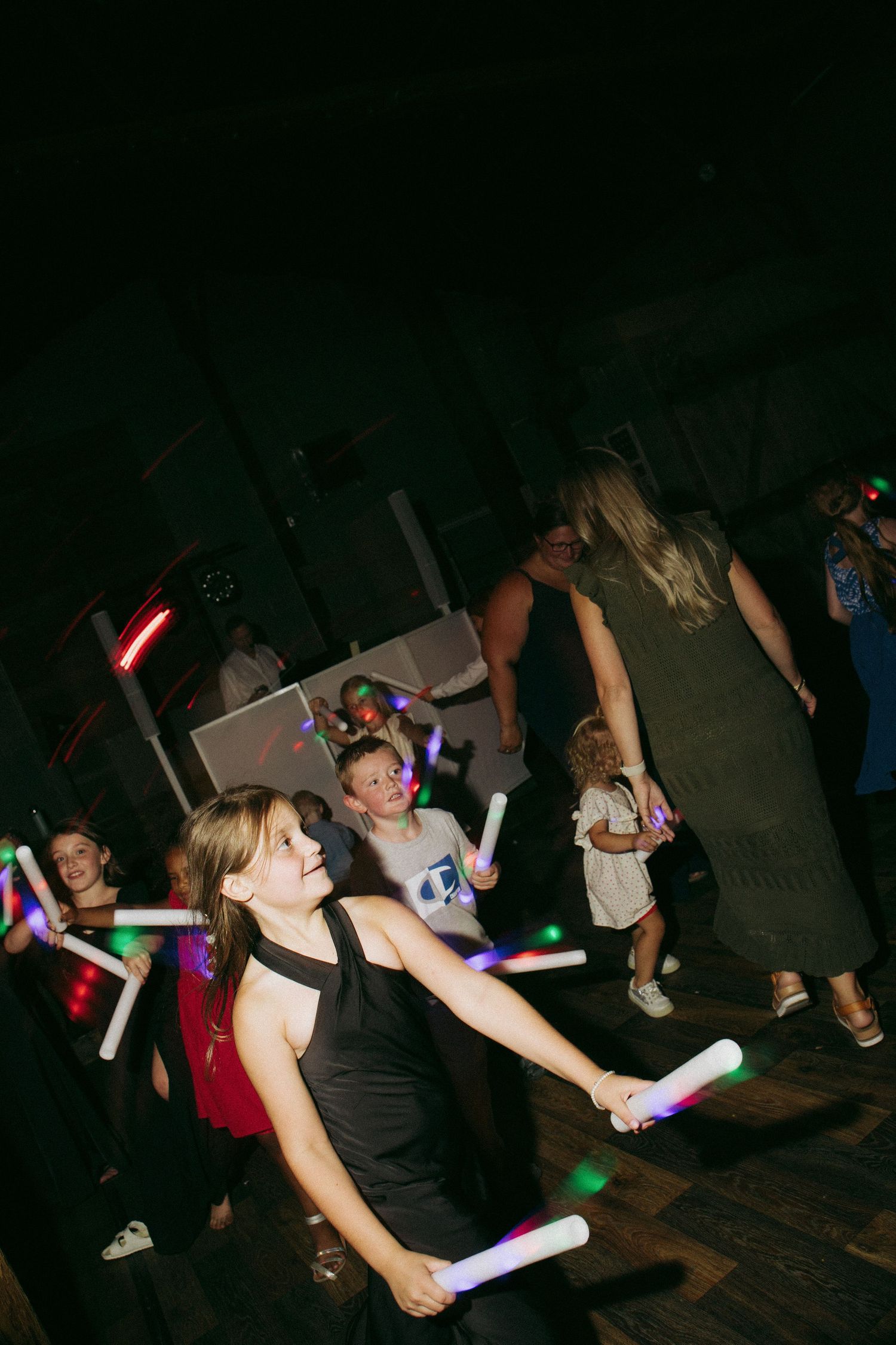 Children and adults dancing with glowing foam sticks at a dark party or wedding reception event.