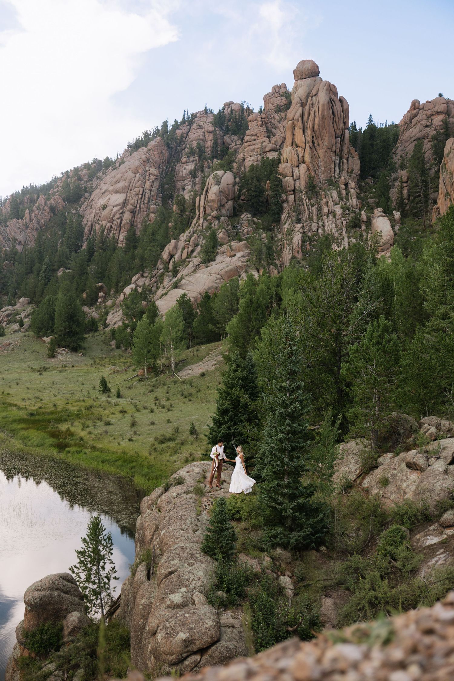 Bride and groom stand on rocky mountain ledge beside a reflective lake surrounded by pine trees and dramatic rock formations.