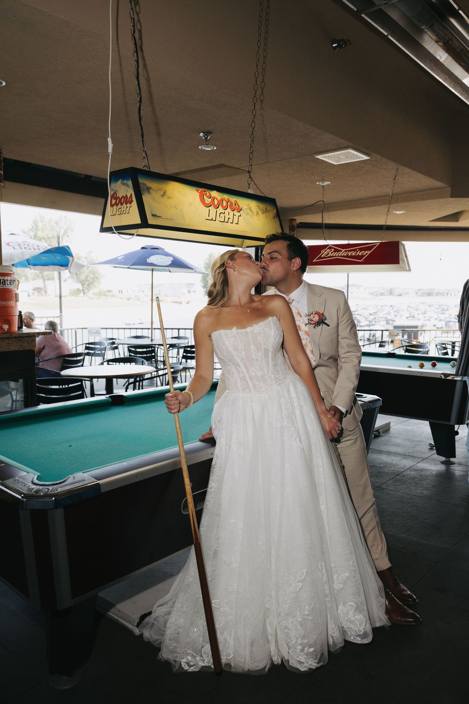 Bride and groom kissing beside a pool table under a Coors Light sign, bride holding a cue stick in a lace wedding gown.