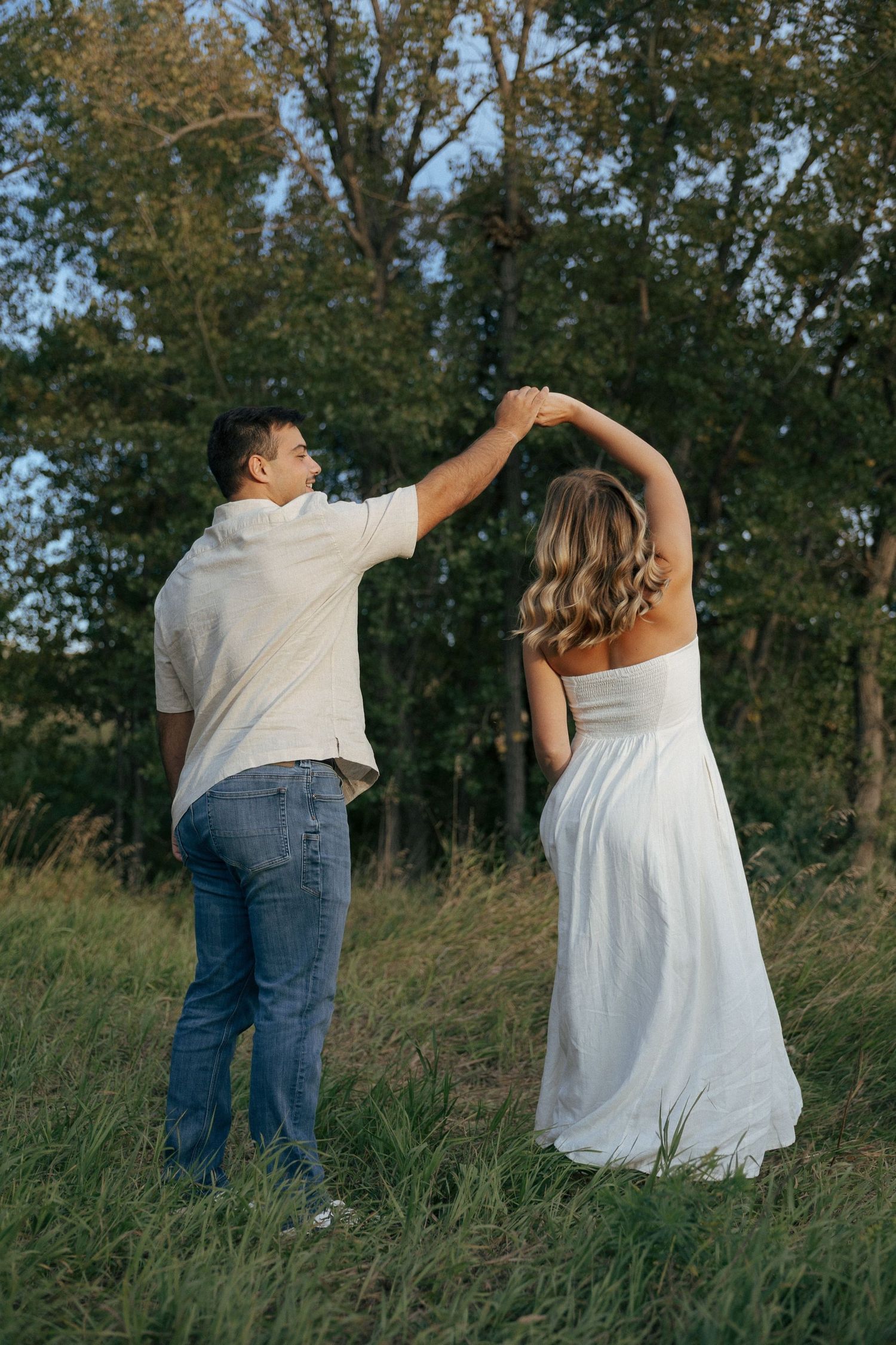 A couple dancing together in a grassy field with trees in the background during a romantic photoshoot.