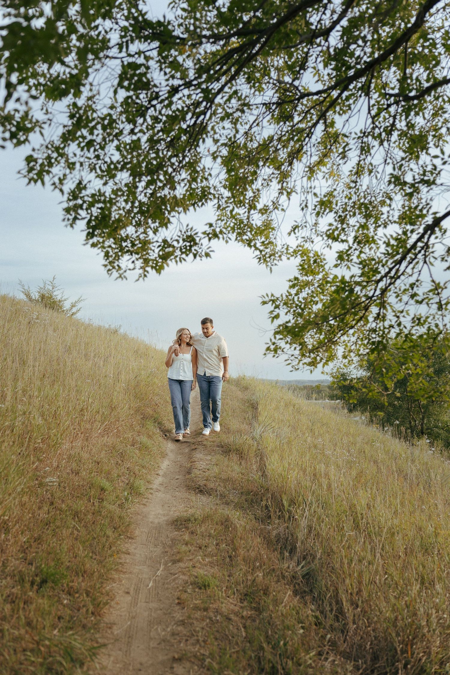 A couple walks hand in hand along a scenic hillside trail under overhanging tree branches.