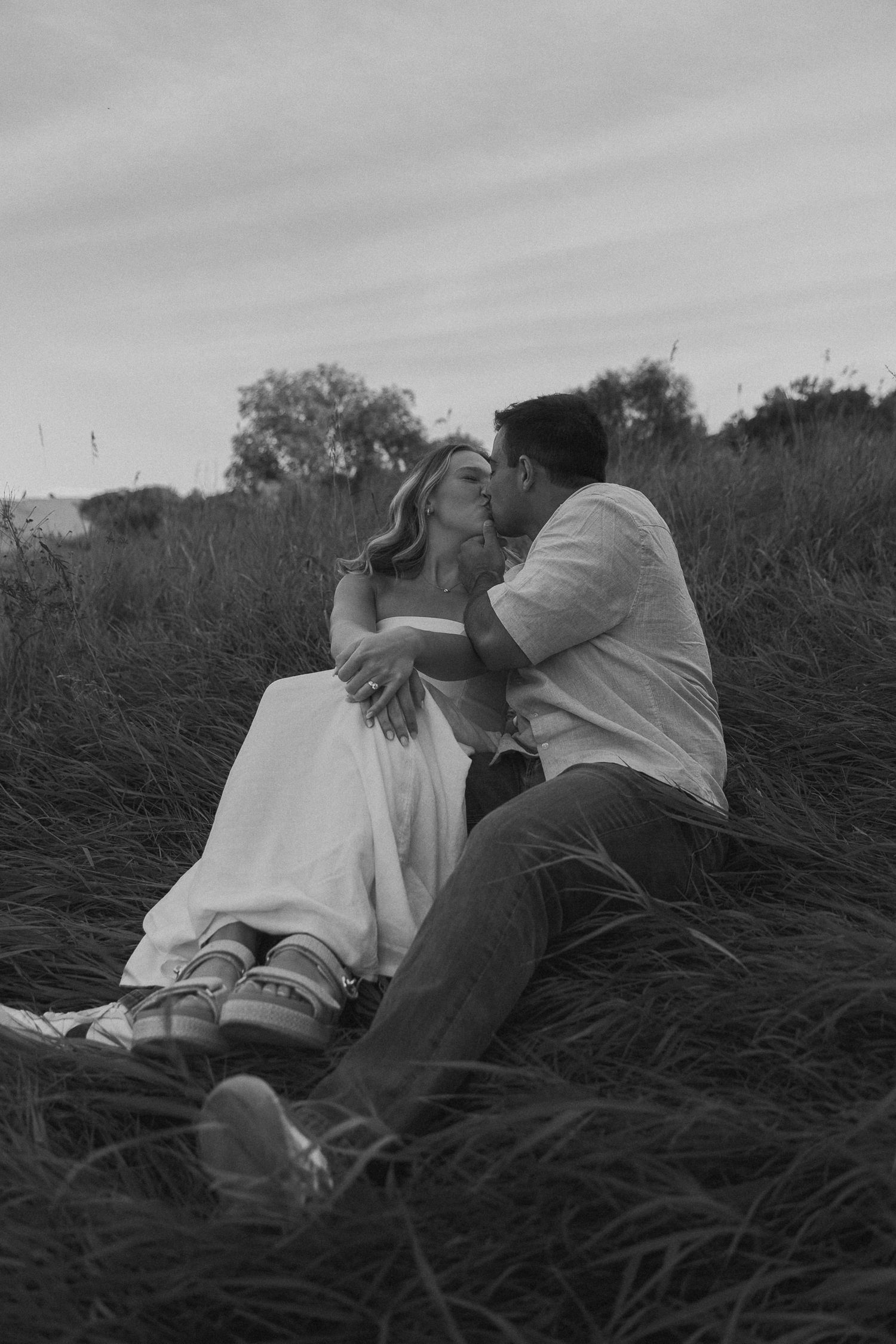 A romantic black and white photo of a couple lying in tall grass during a sunset photoshoot.