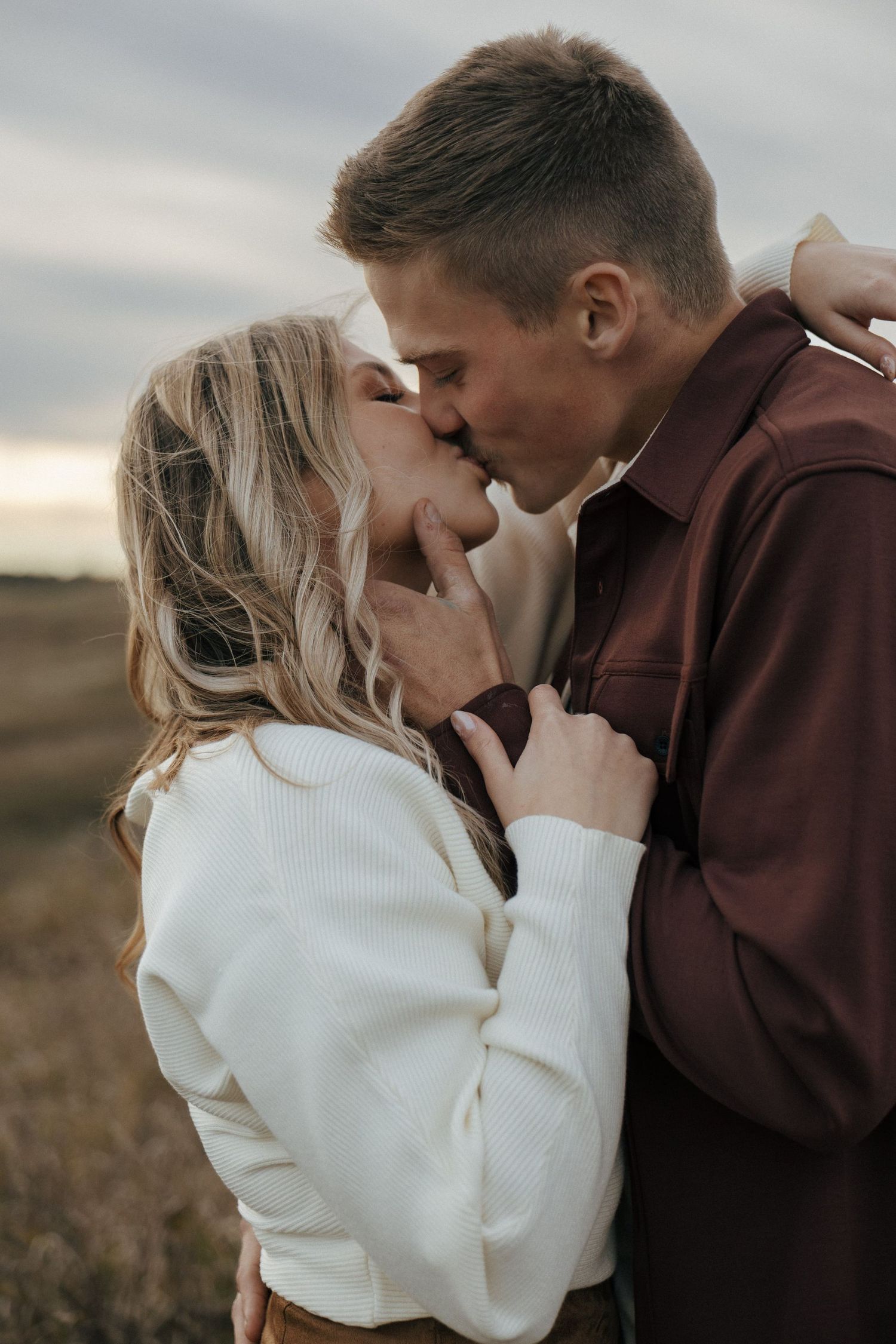 A tender moment between a couple kissing in a field, with her wearing white and him in burgundy.