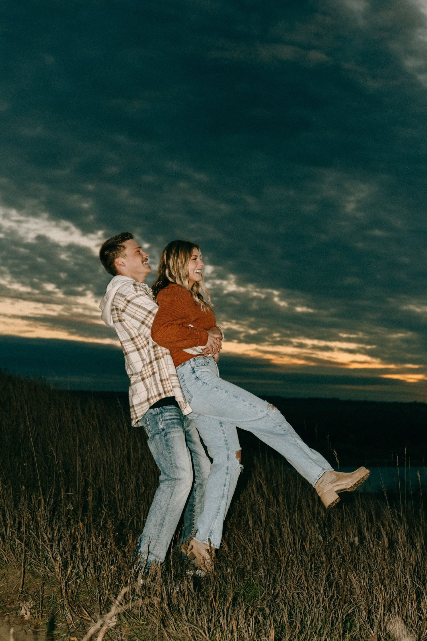 Playful couple poses in field during sunset with dramatic cloudy sky.