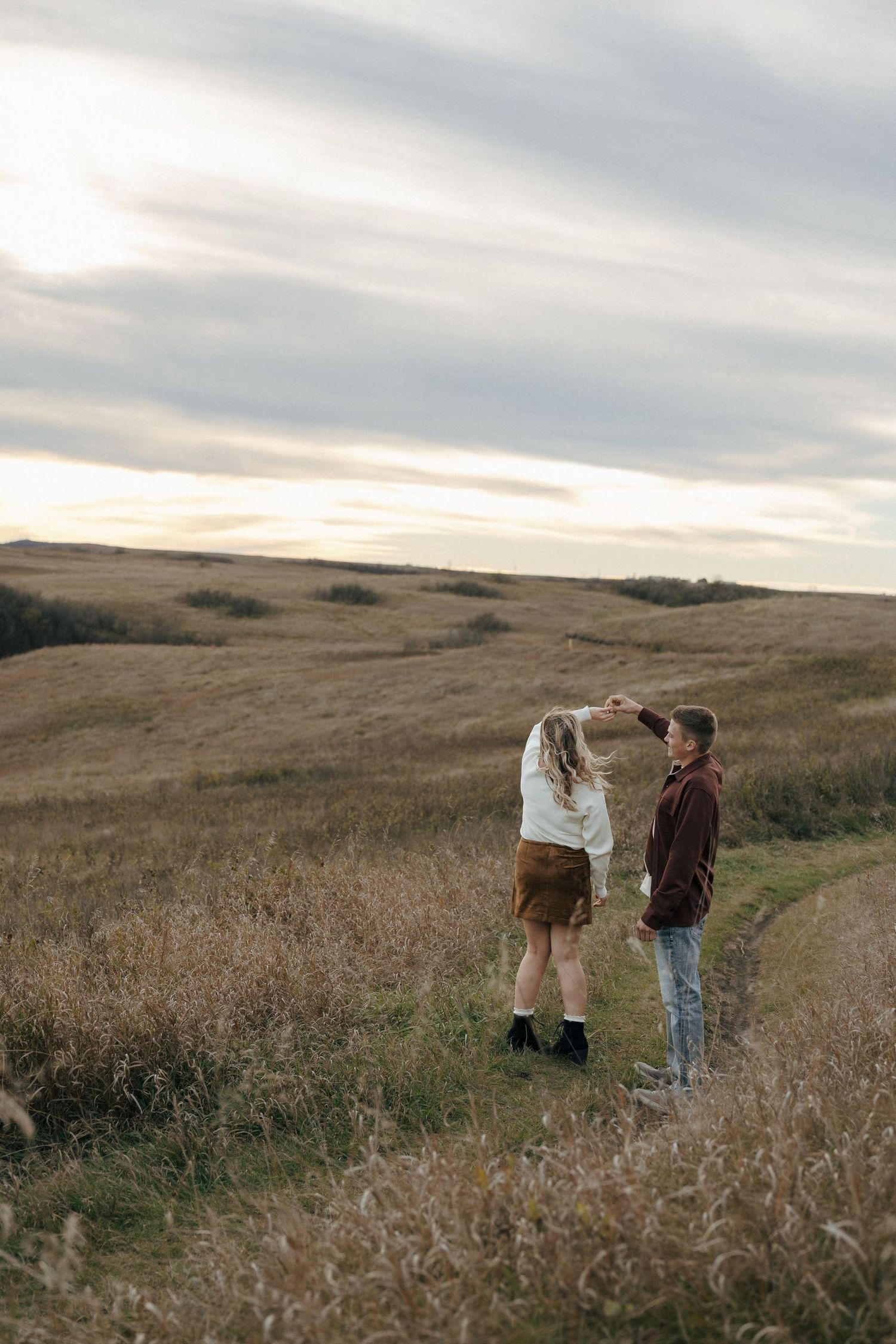 A couple walks through a golden grass field at sunset against rolling hills in the distance.