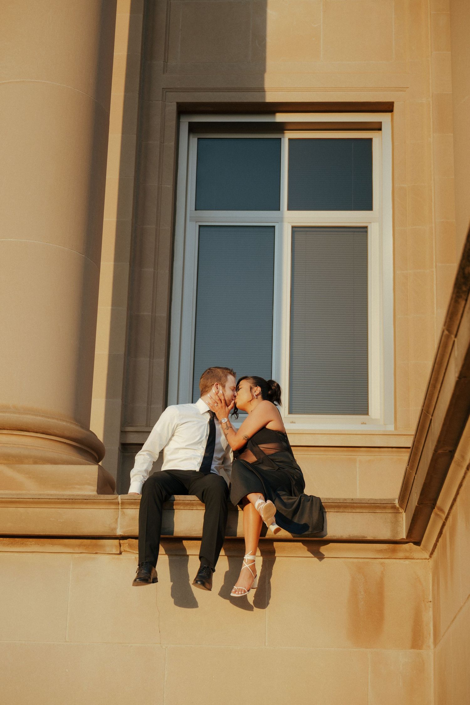 A couple shares an intimate moment on a building ledge in warm sunlight.
